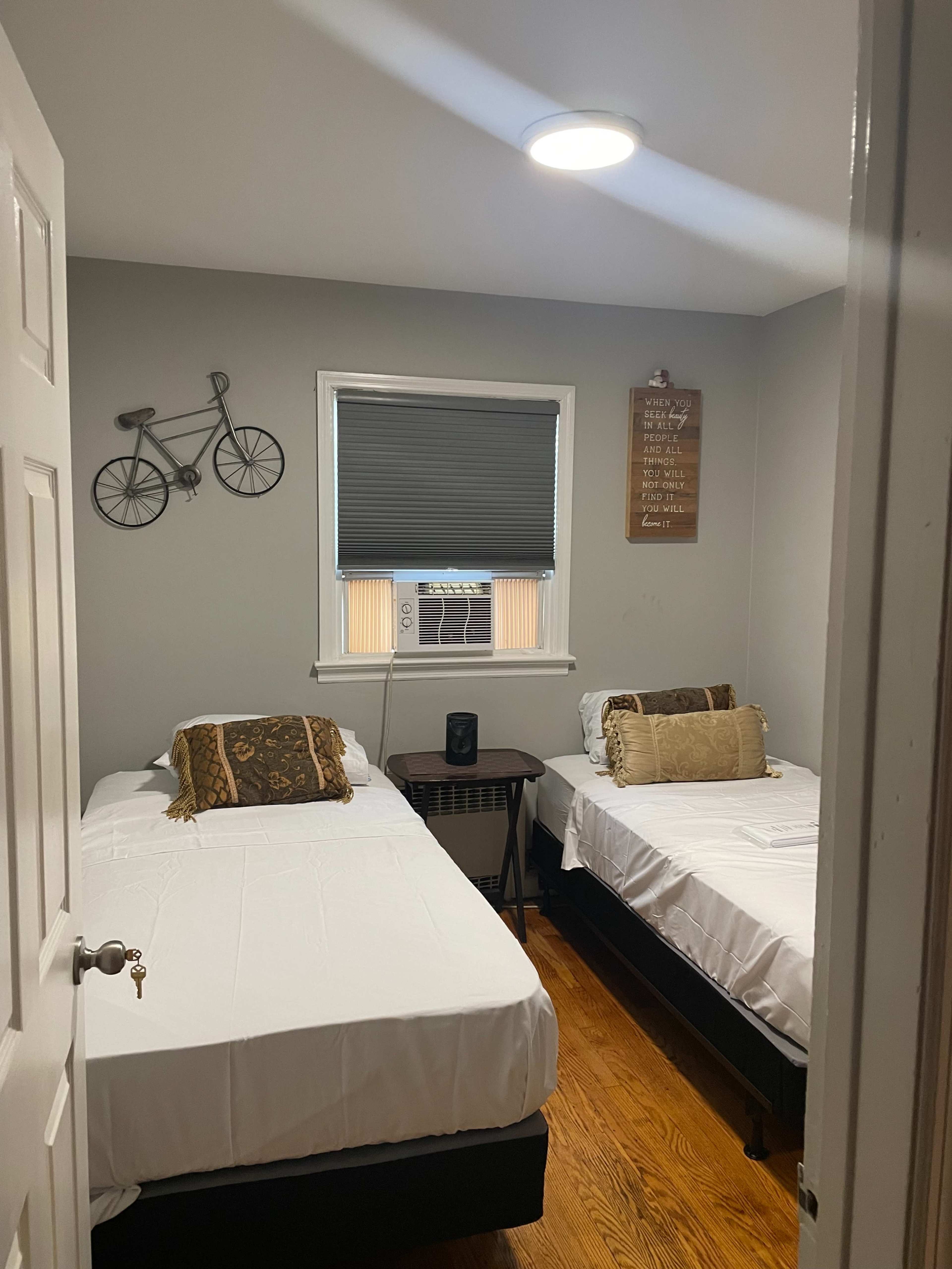 The image shows a small bedroom with two single beds, a side table, and decorative items on the walls.