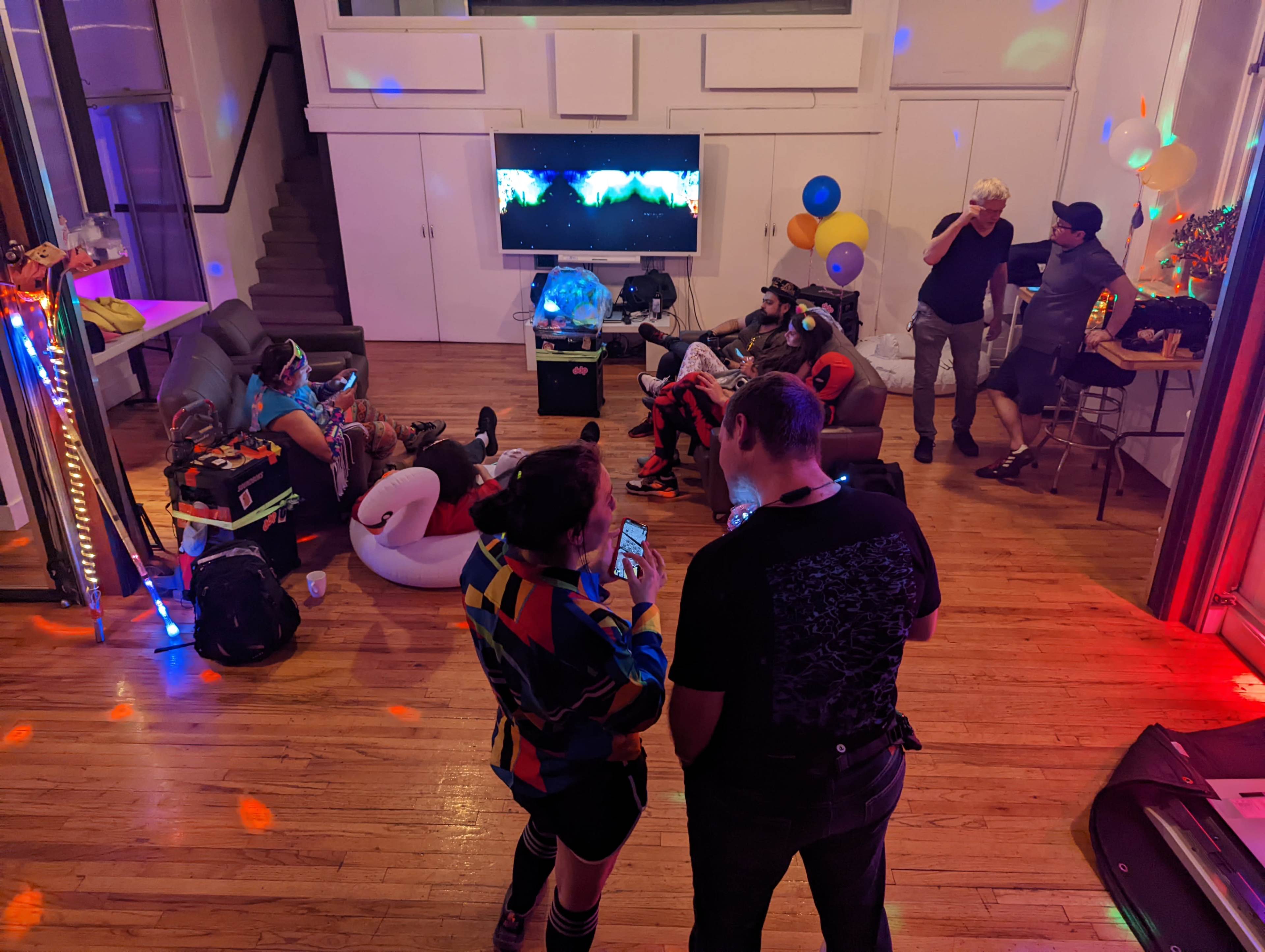 The image shows a lively indoor party scene with colorful lights, guests lounging on furniture, and balloons decorating the space.