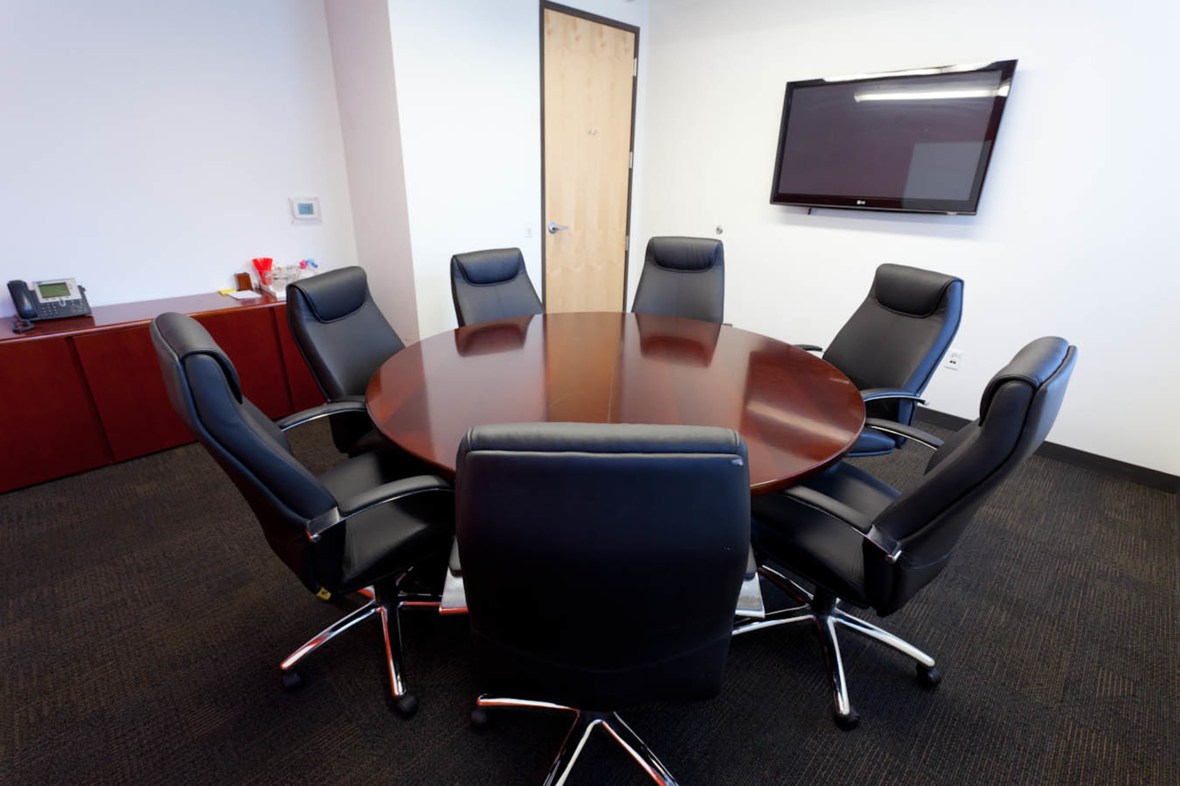 A round wooden conference table surrounded by seven black leather chairs in a modern meeting room.