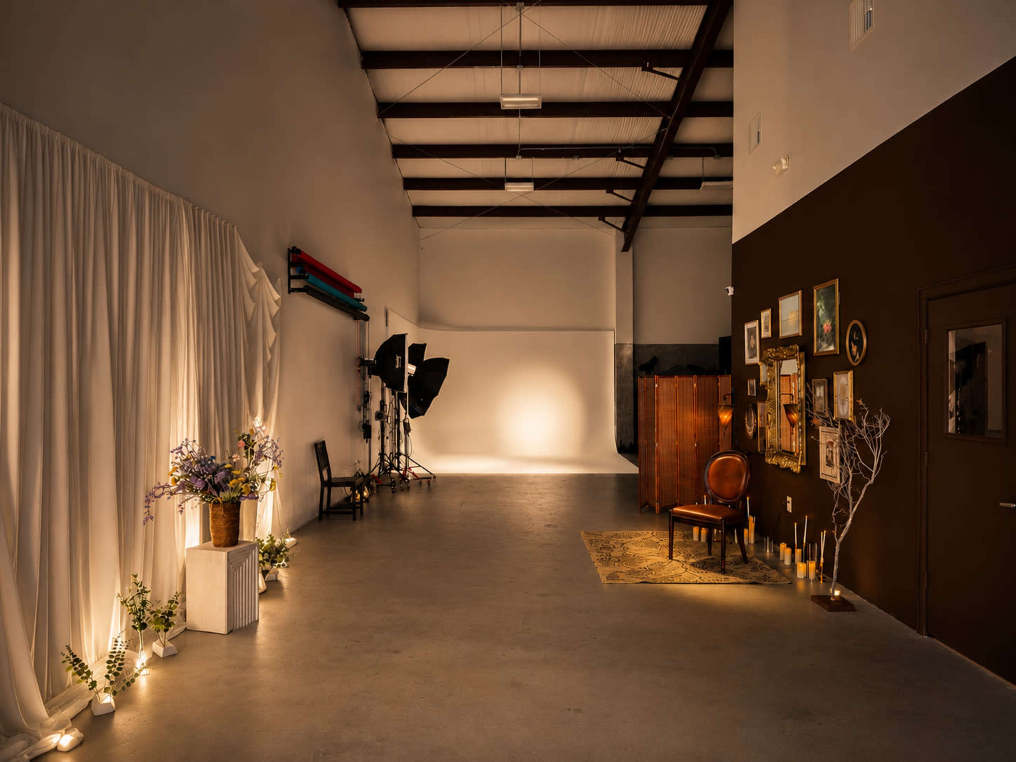 The image shows a spacious photography studio with a draped backdrop, lighting equipment, and various decorative elements, including plants and framed artwork.