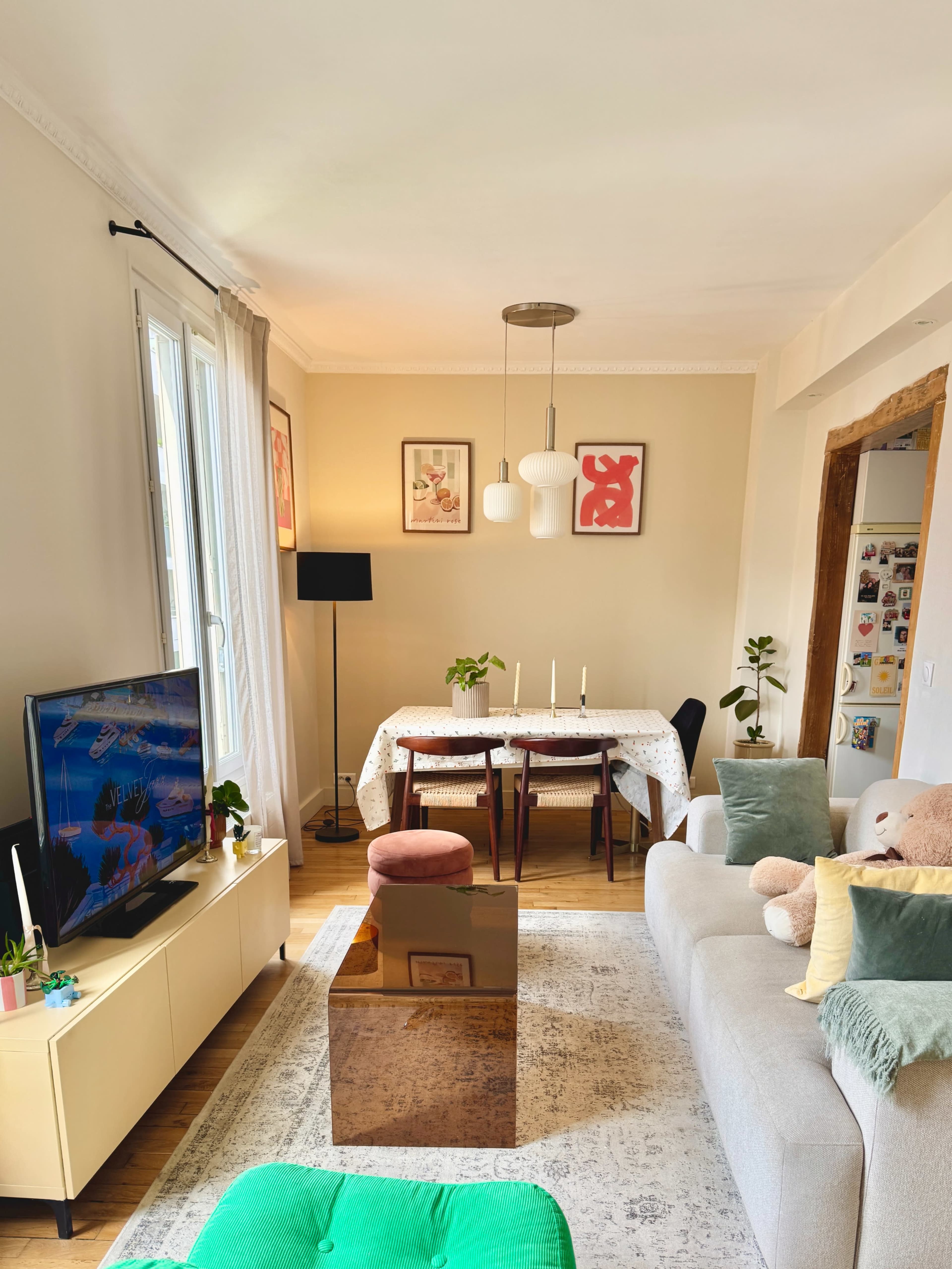 The image shows a living room with a television, a dining table set for four, and a gray sofa with decorative pillows, illuminated by natural light from a window.