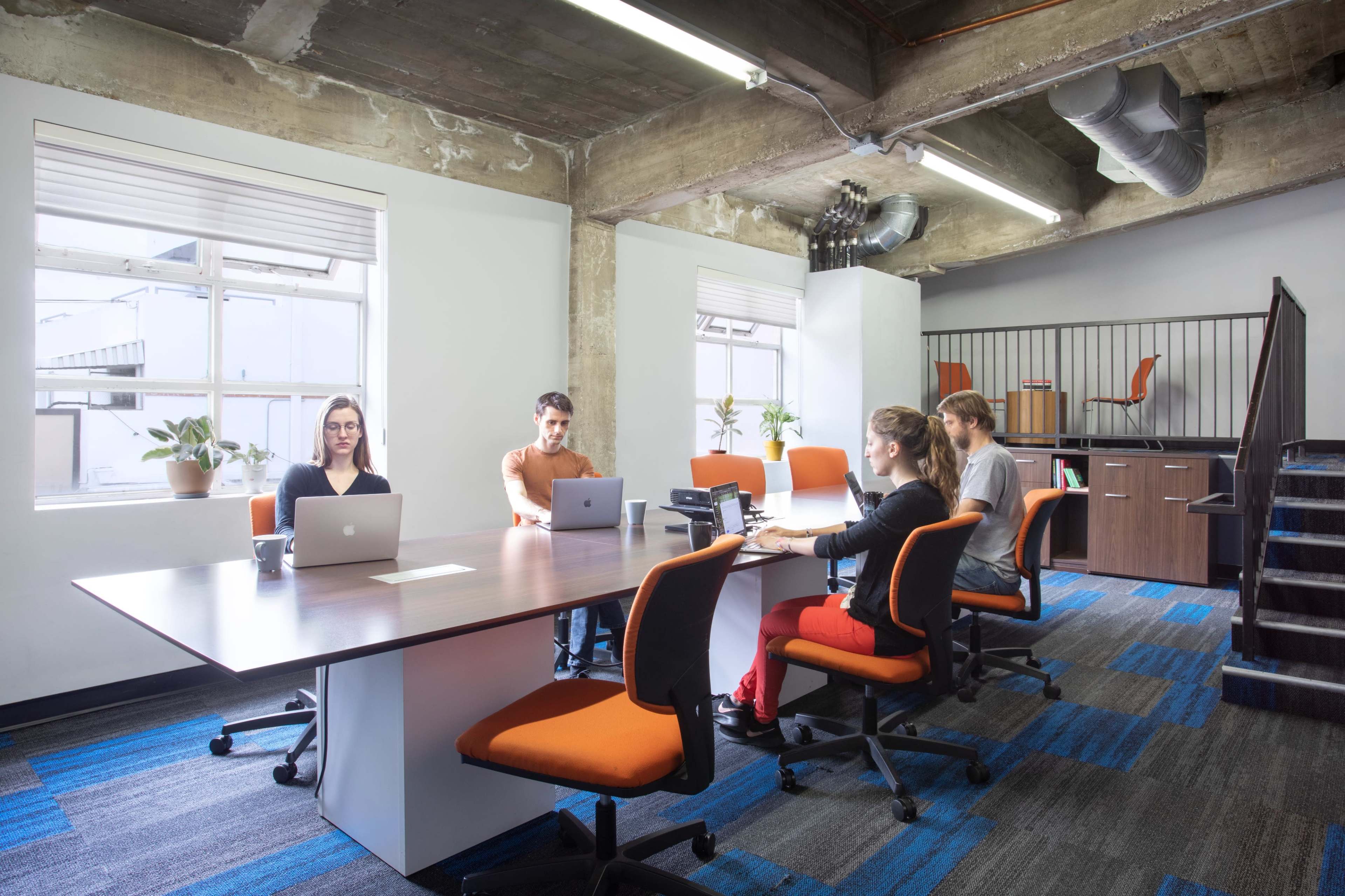 A group of four people work at a large table in a modern office with concrete walls and bright orange chairs.