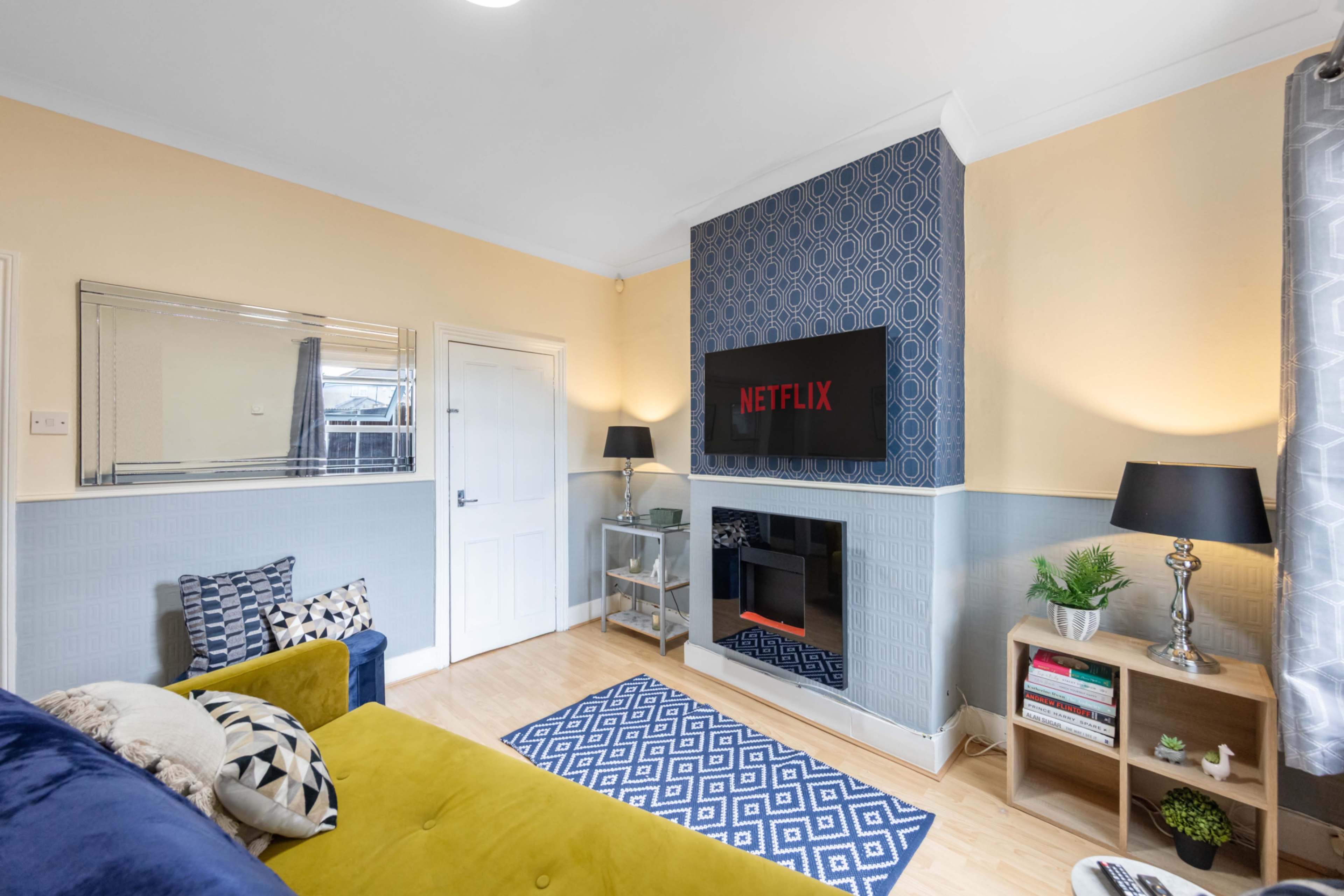 The living room features a blue and yellow color scheme with a television mounted on a patterned wall, a fireplace, and a patterned rug.