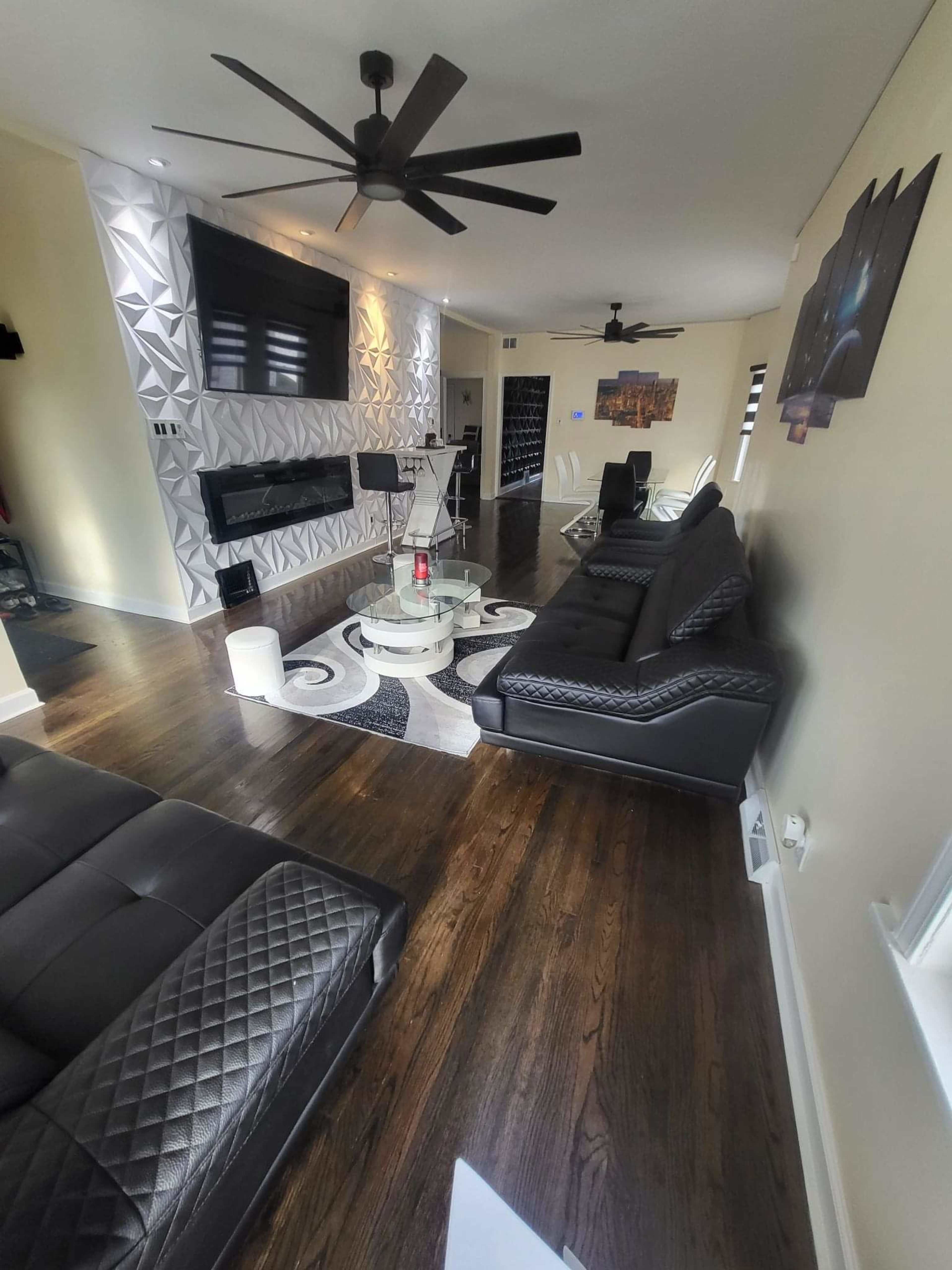 The image depicts a modern living room with black leather furniture, a glass coffee table, and decorative wall features, all arranged in an open layout.