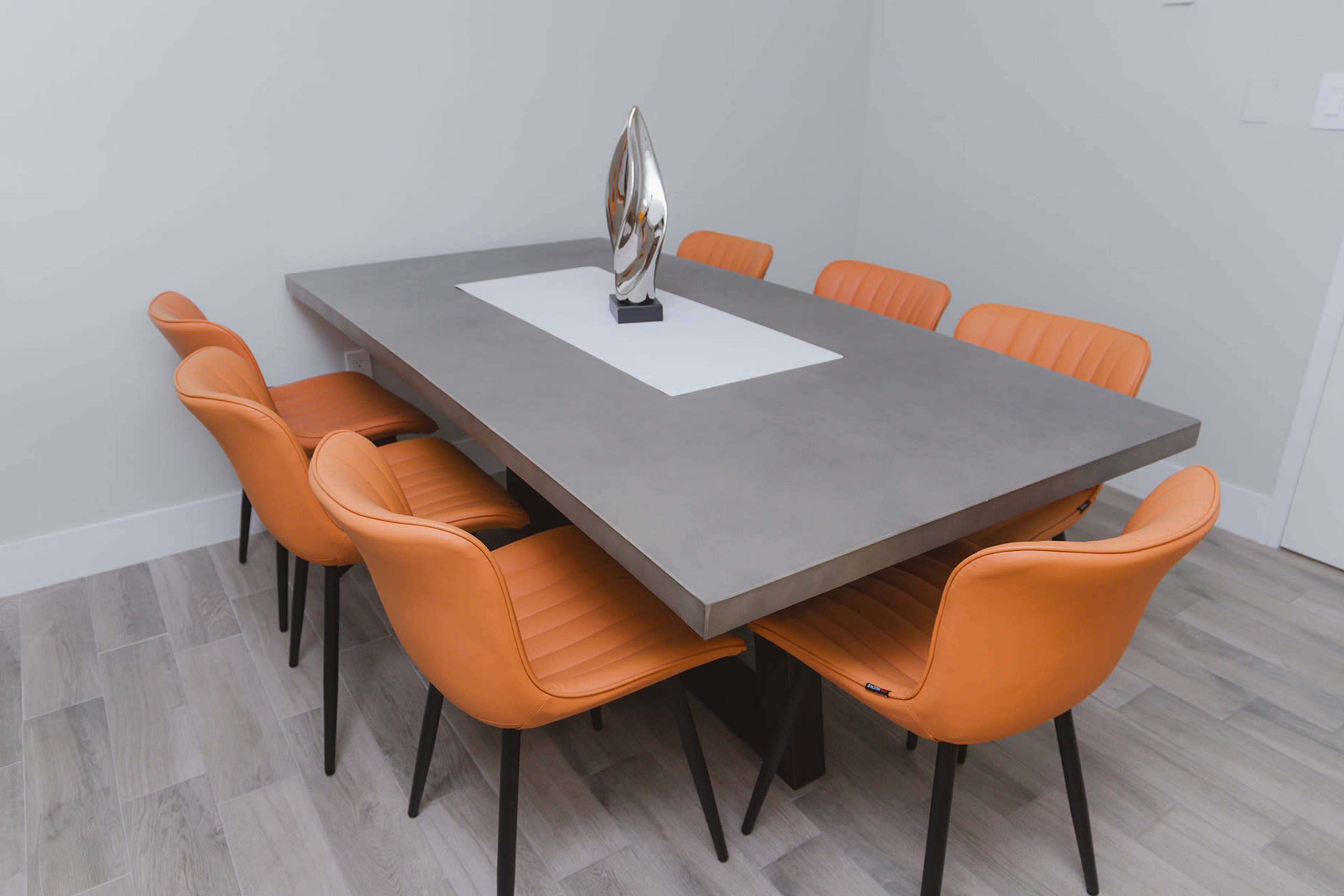 A rectangular concrete dining table is surrounded by eight orange chairs in a minimalist room.