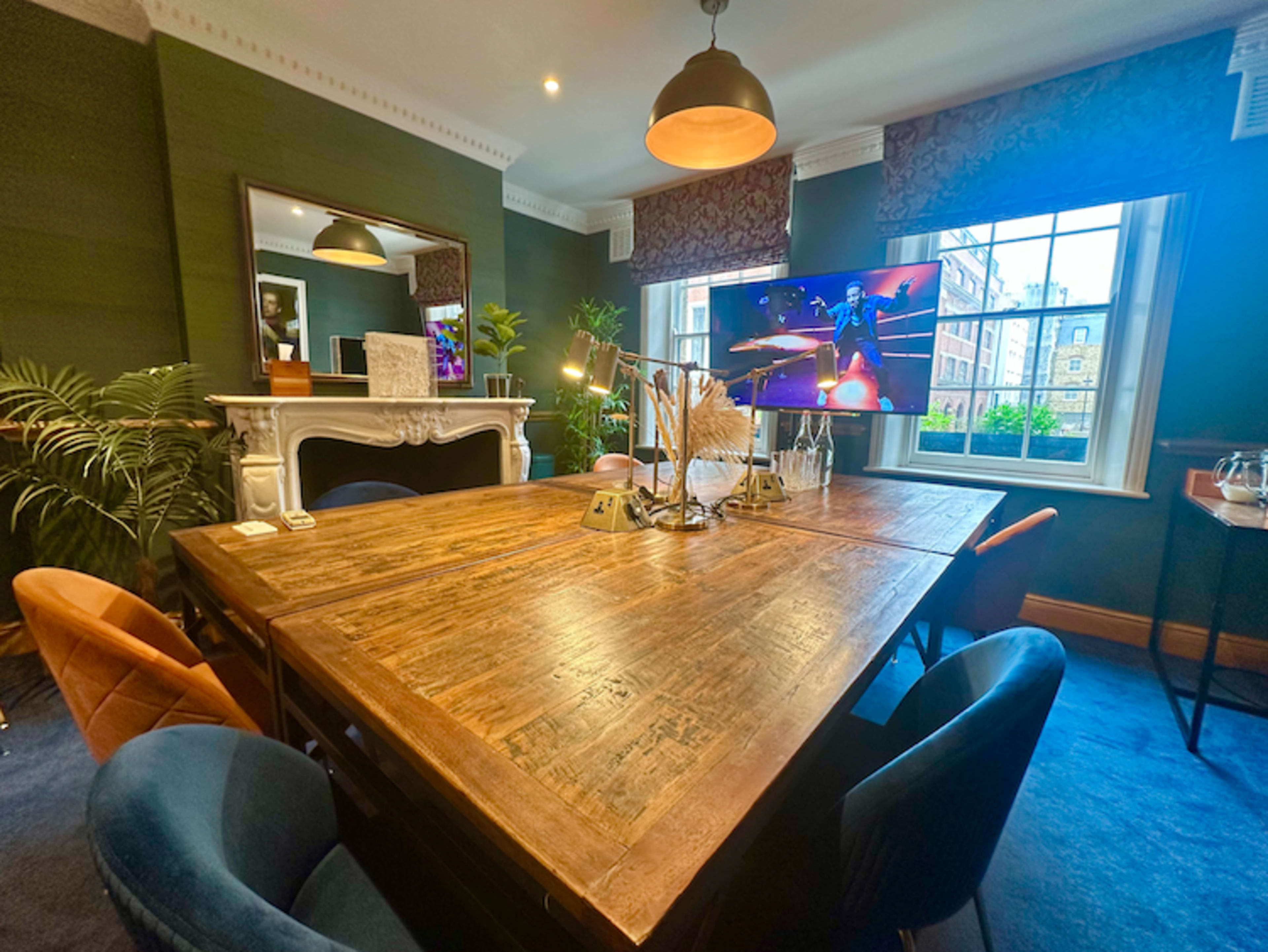 A large wooden table is centered in a stylish meeting room, surrounded by upholstered chairs and featuring a television mounted on the wall and a decorative fireplace.