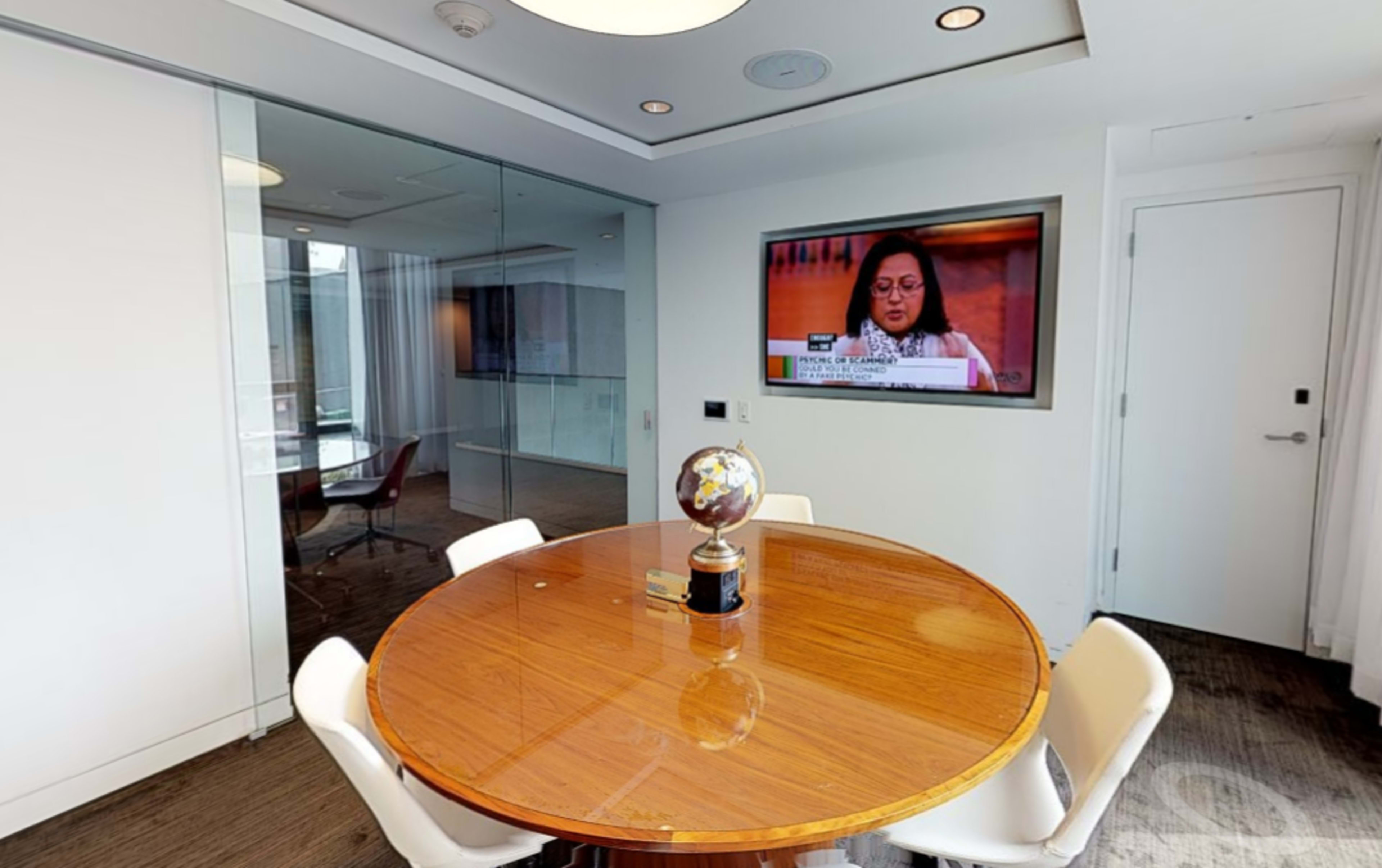 A round wooden table with four white chairs is positioned in a meeting room, featuring a television displaying a news program on the wall.