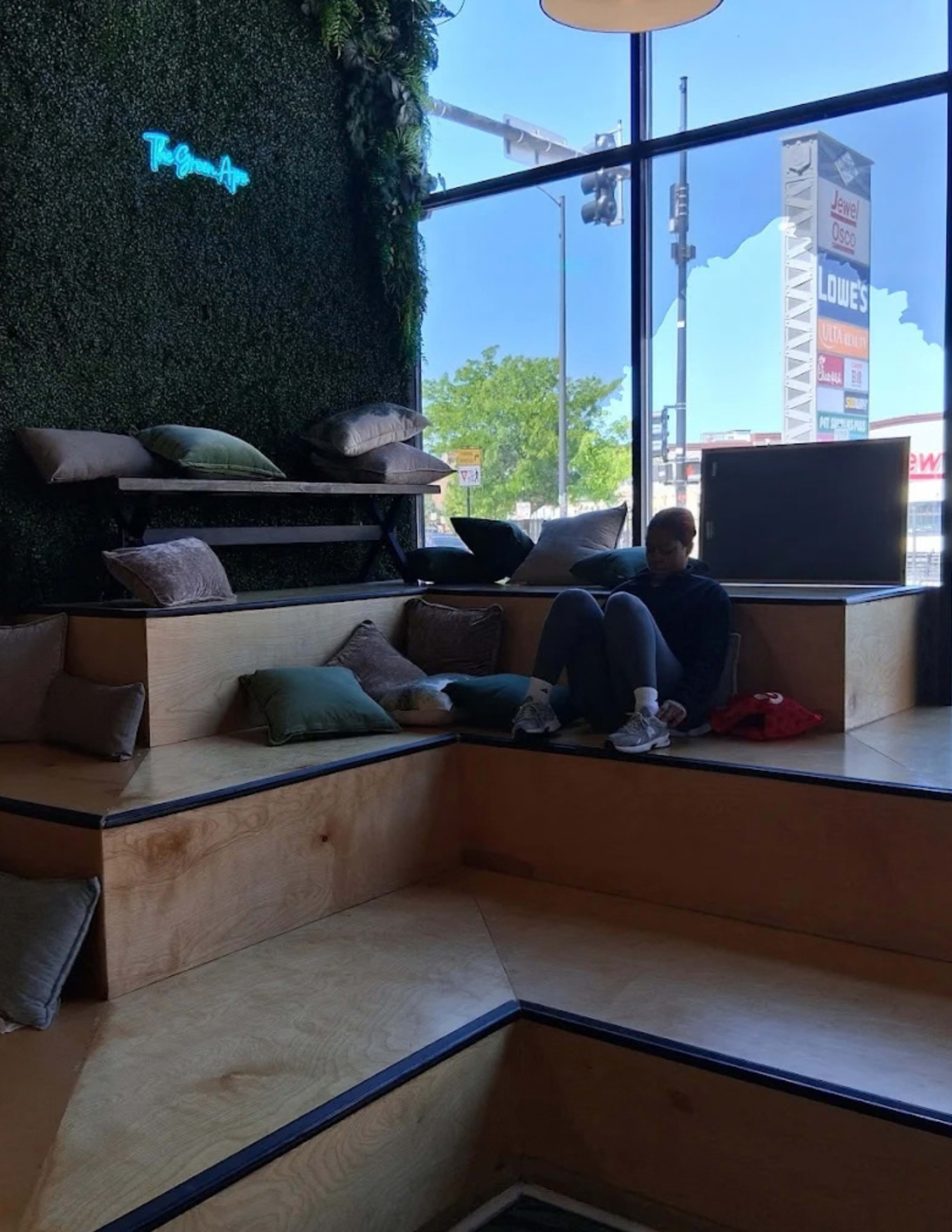 A person is sitting on a wooden seating area with cushions, in a space that features greenery and large windows.