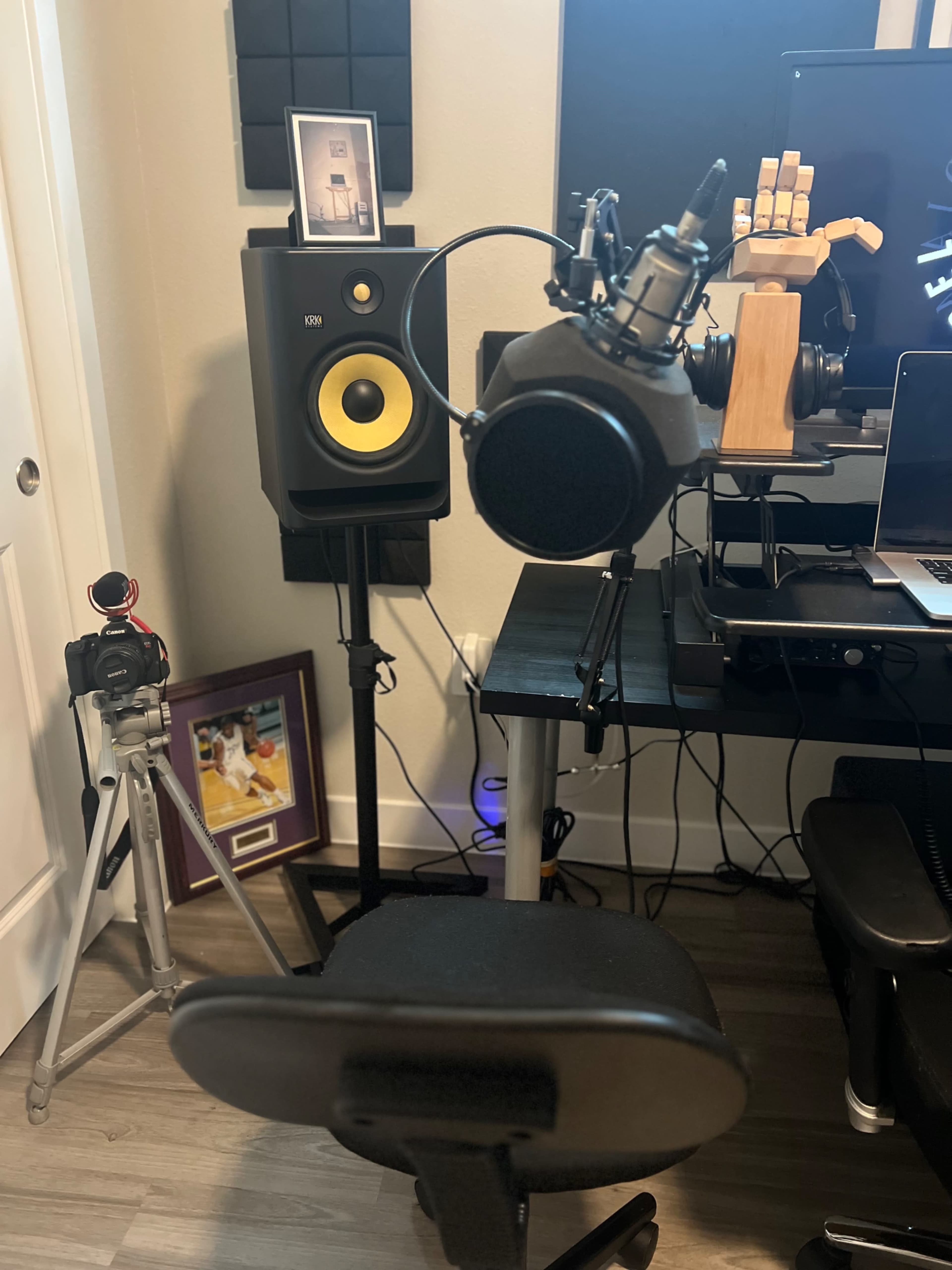A home studio setup with a microphone mounted on a stand, a speaker on a stand, a camera on a tripod, and a computer workstation.