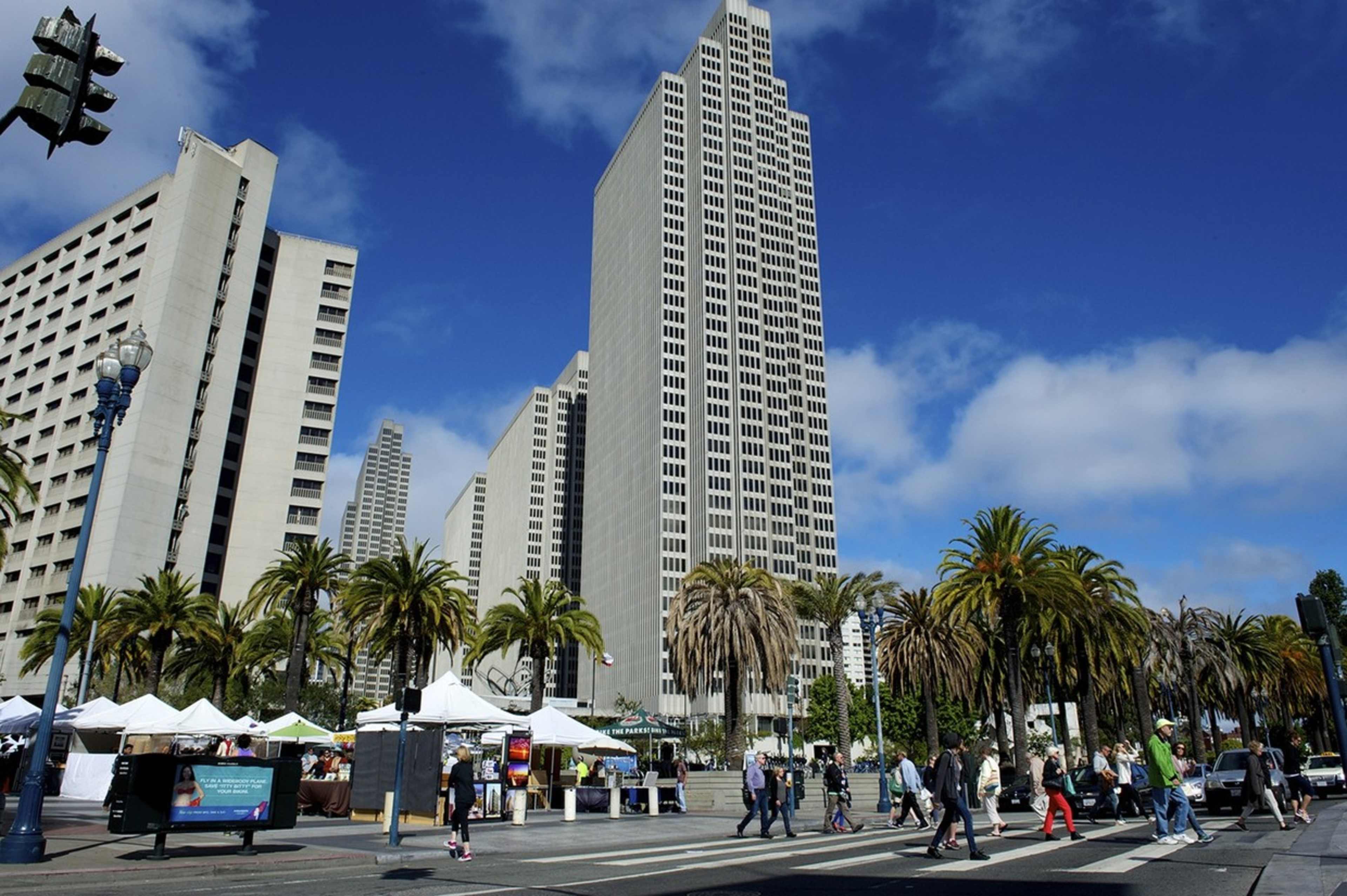 A bustling city street with people walking past palm trees and vendors near tall, modern skyscrapers under a partly cloudy sky.