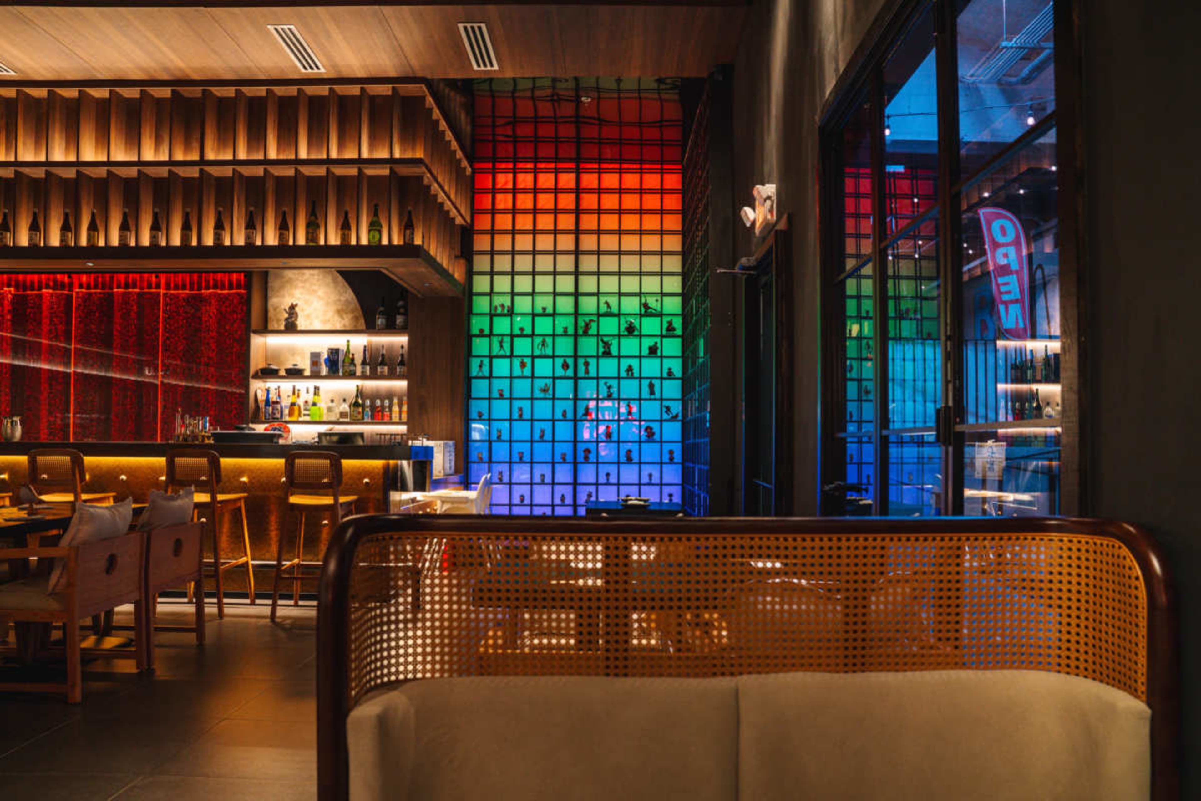 The image shows a modern restaurant interior with a colorful illuminated wall and a bar area in the background.