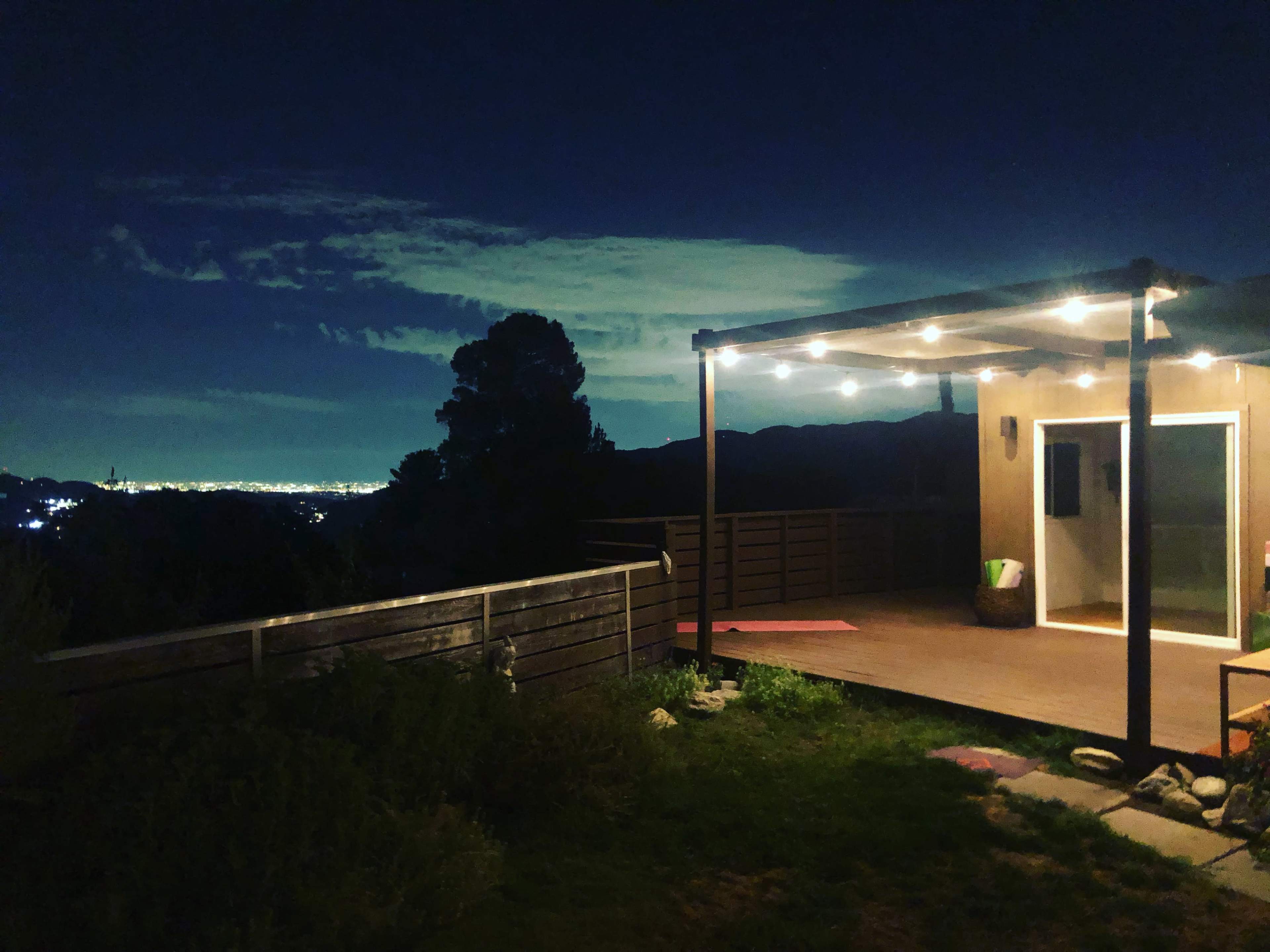 A deck with string lights overlooks a city skyline at night, surrounded by trees and plants.