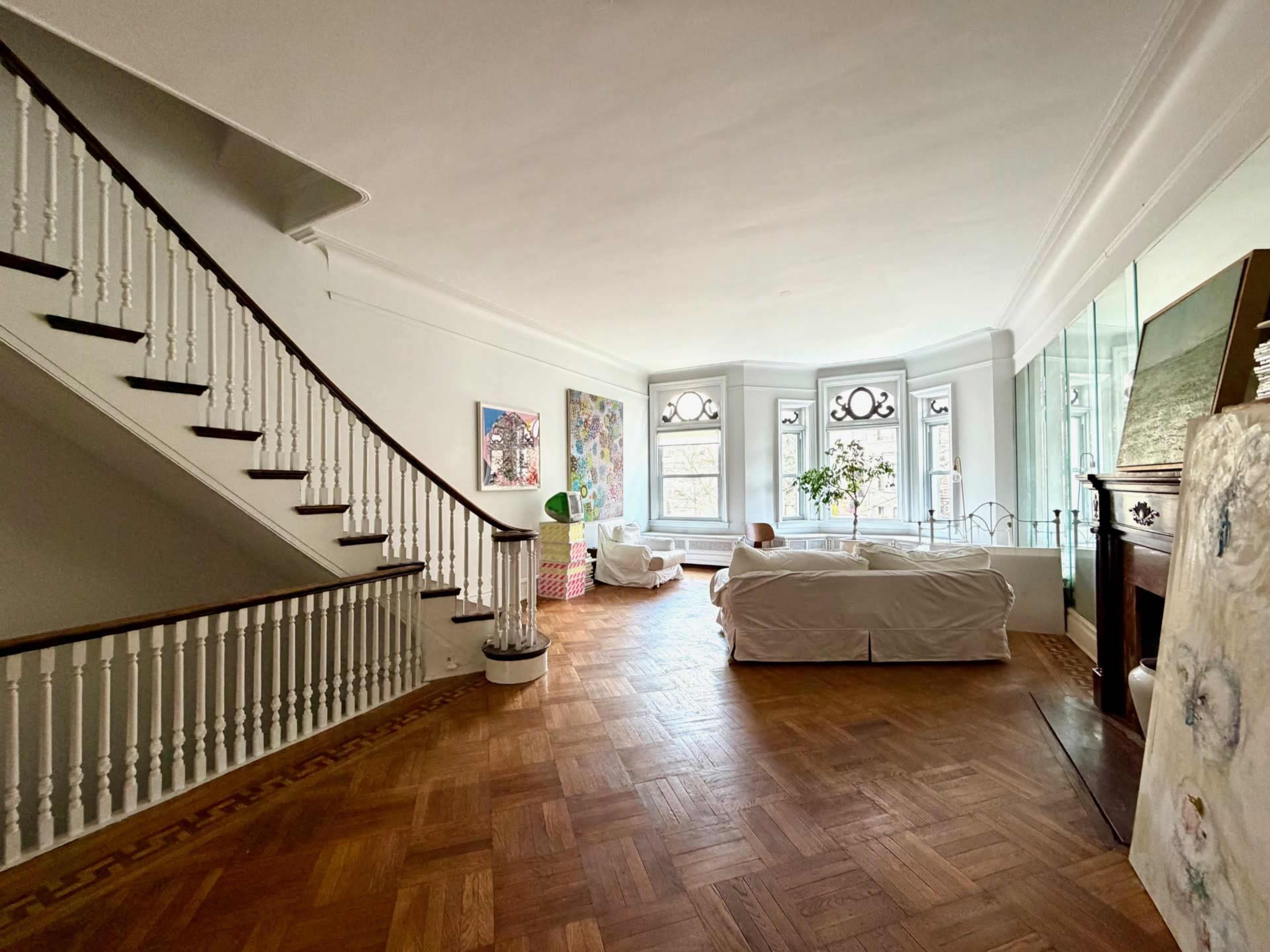 A spacious interior features a staircase on the left, a large window with decorative detailing, and a white couch positioned in the center of the room.