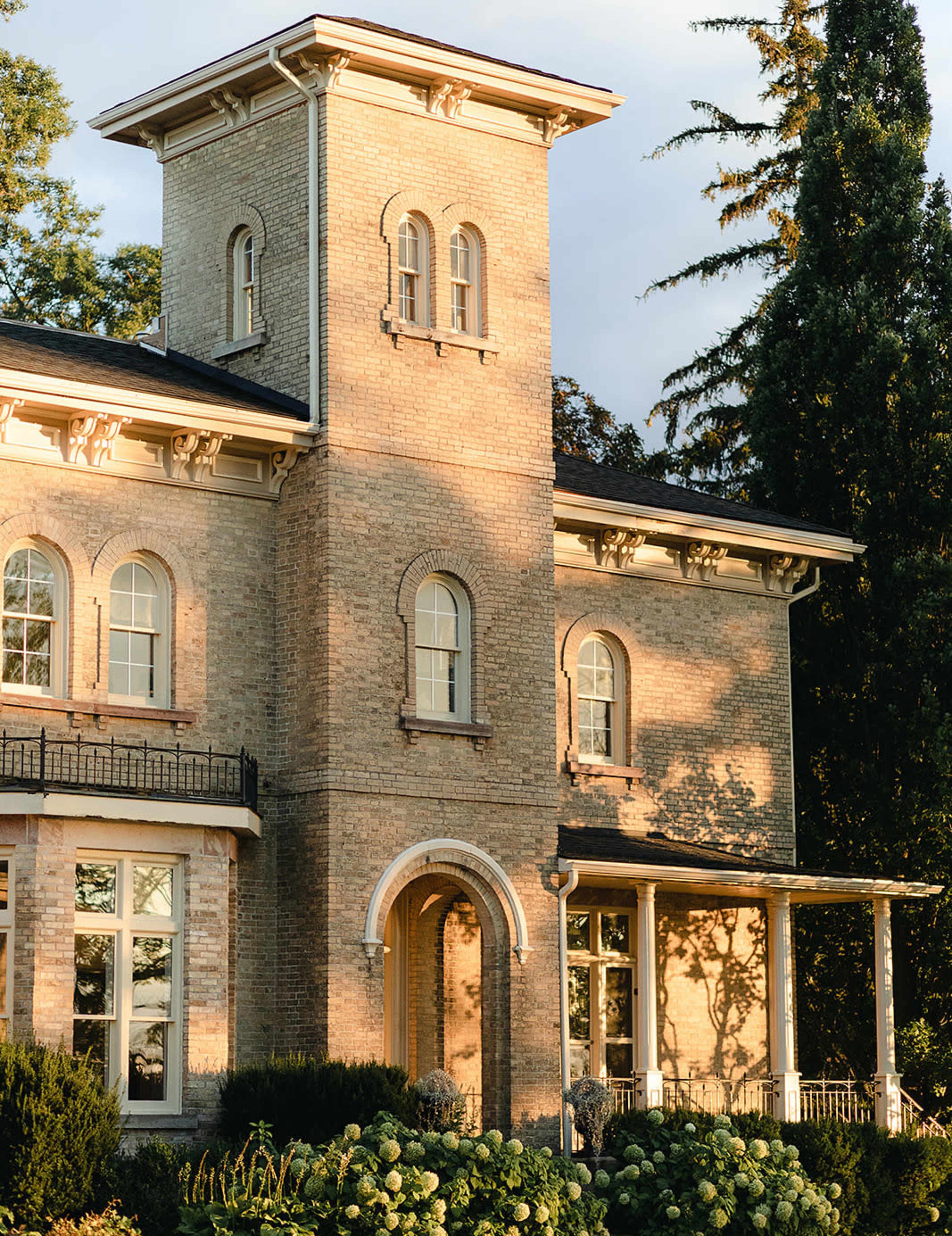 The image depicts a two-story brick house with large windows, a cylindrical tower, and a front porch surrounded by greenery.