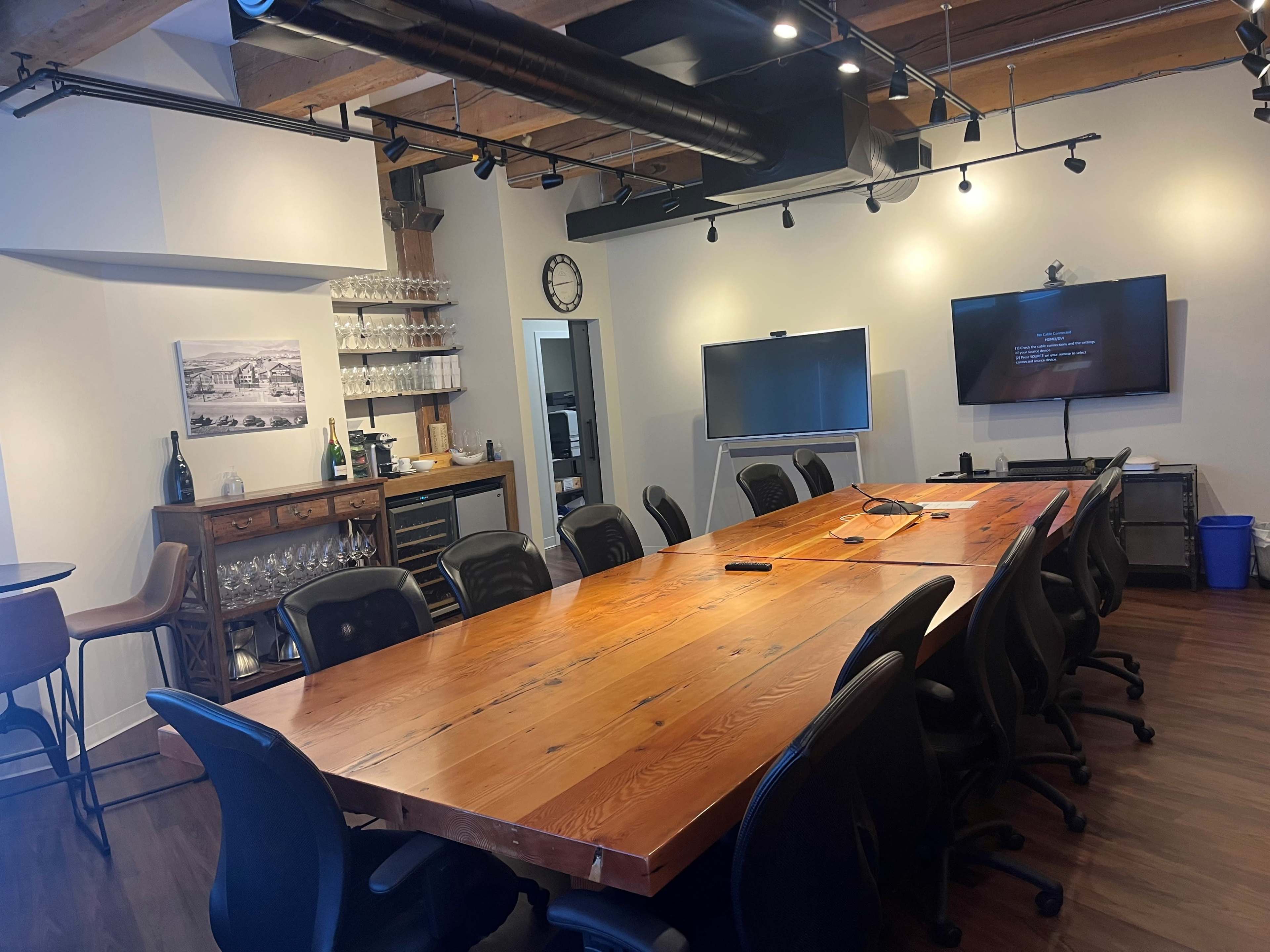 A conference room with a large wooden table surrounded by black chairs, a wall-mounted screen, a bar area with glassware, and several light fixtures.