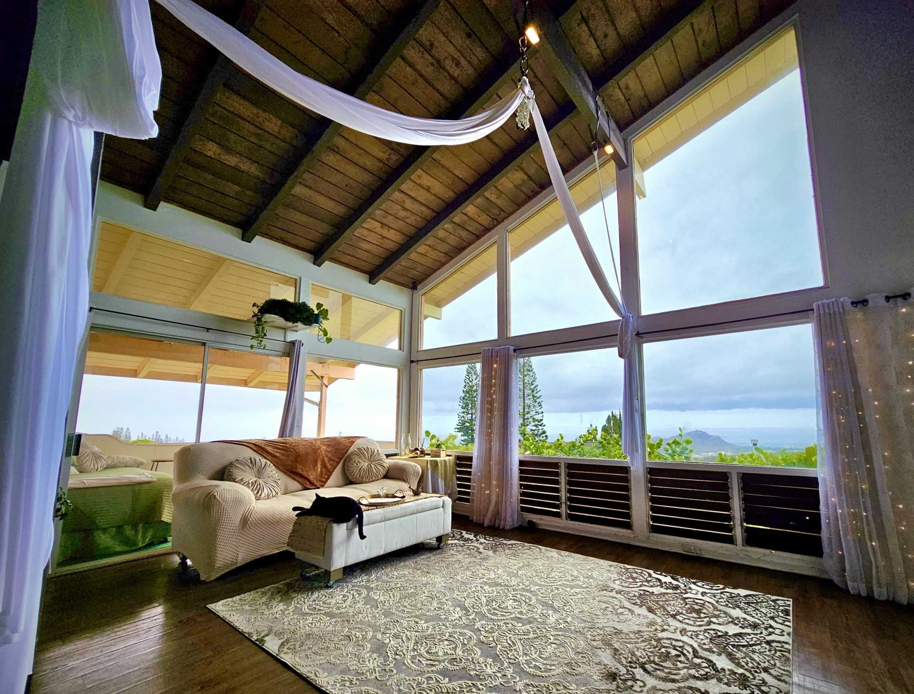 The spacious room features large windows with a view of the outdoors, a comfortable couch, and a decorative rug on the wooden floor.