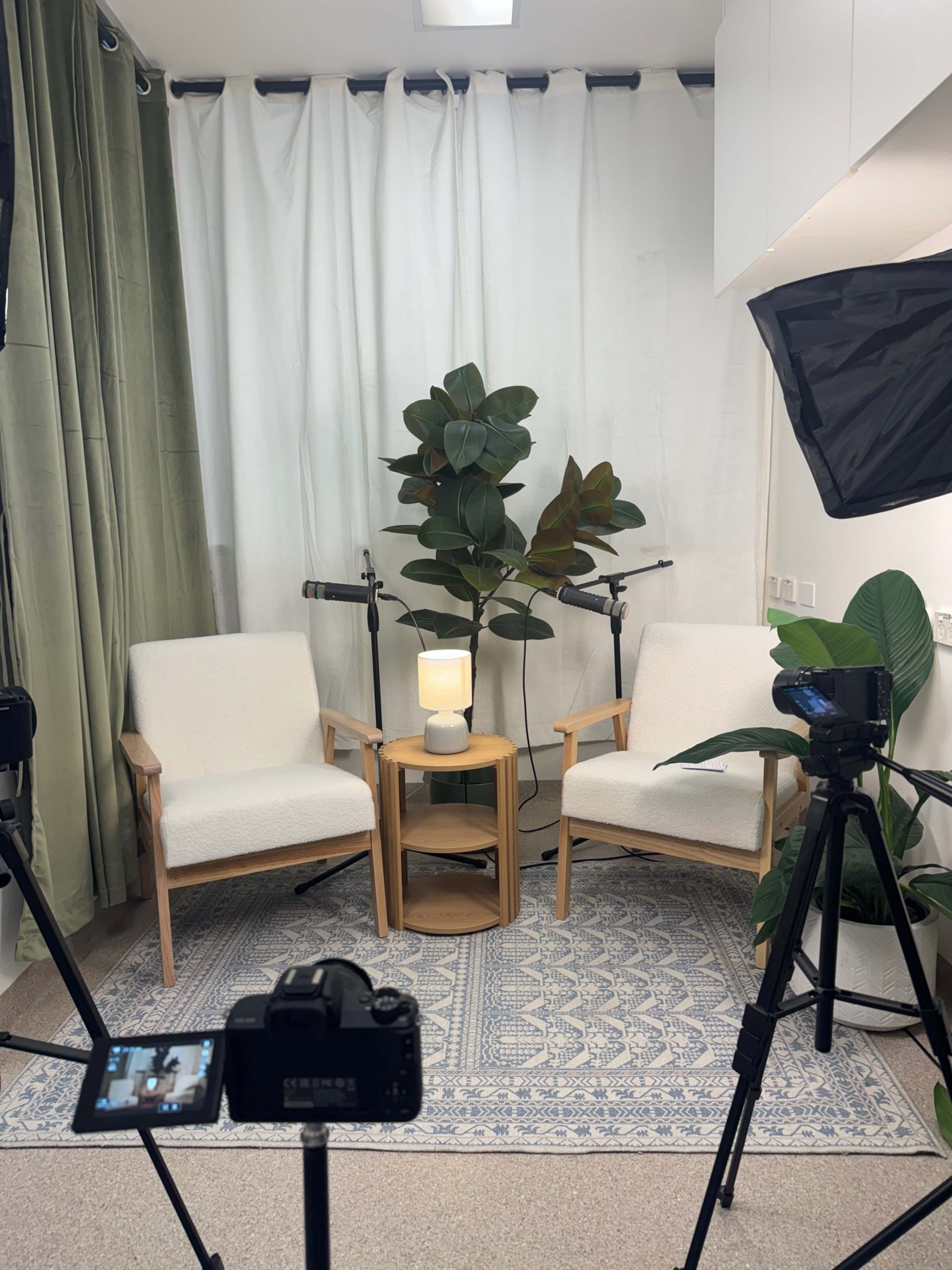 The image shows a small recording setup featuring two white chairs, a round wooden table with a lamp, and a decorative plant, all positioned in front of a white backdrop.