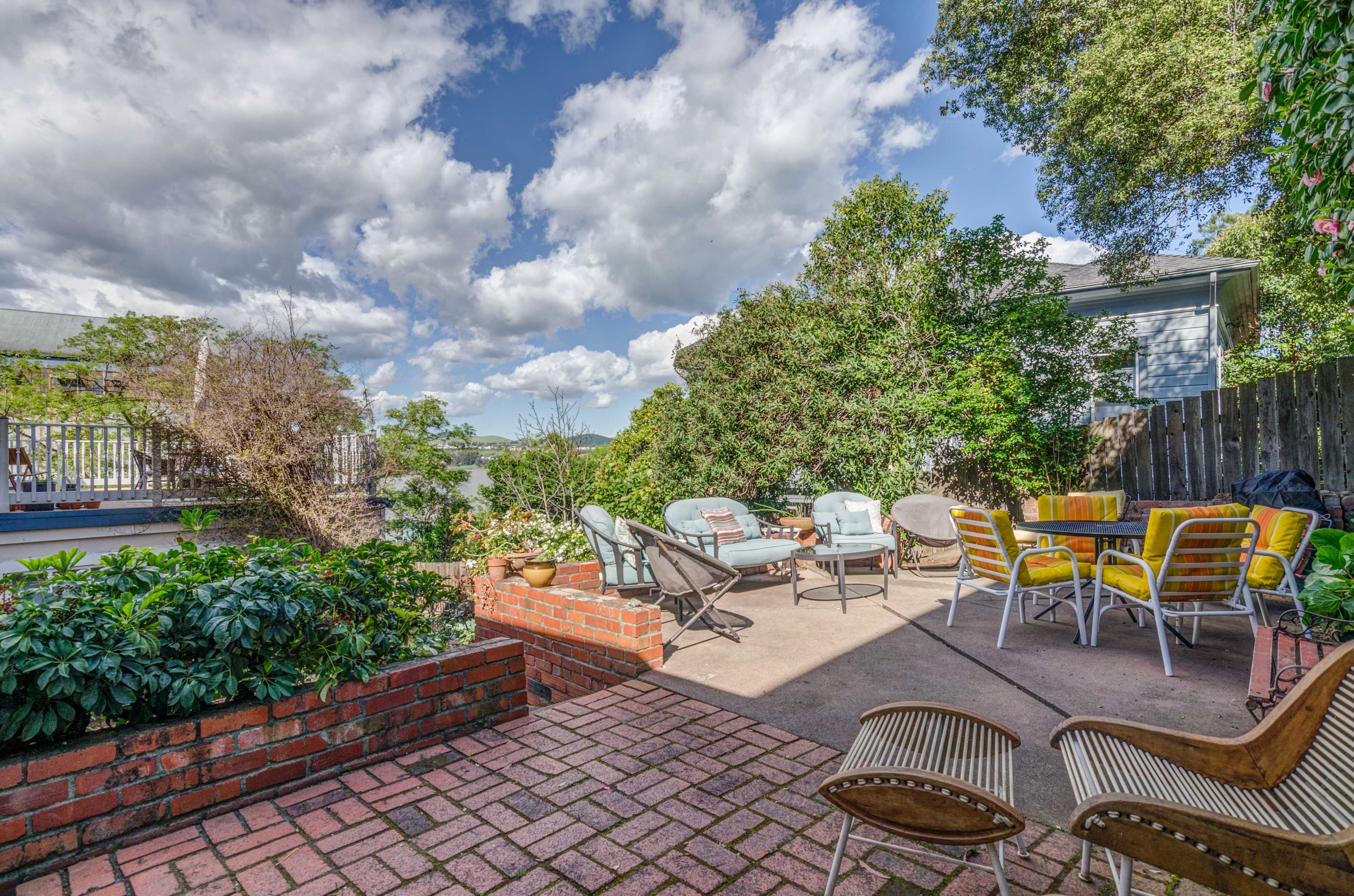 A patio area with brick flooring, surrounded by greenery and featuring seating arrangements of metal chairs and a table.