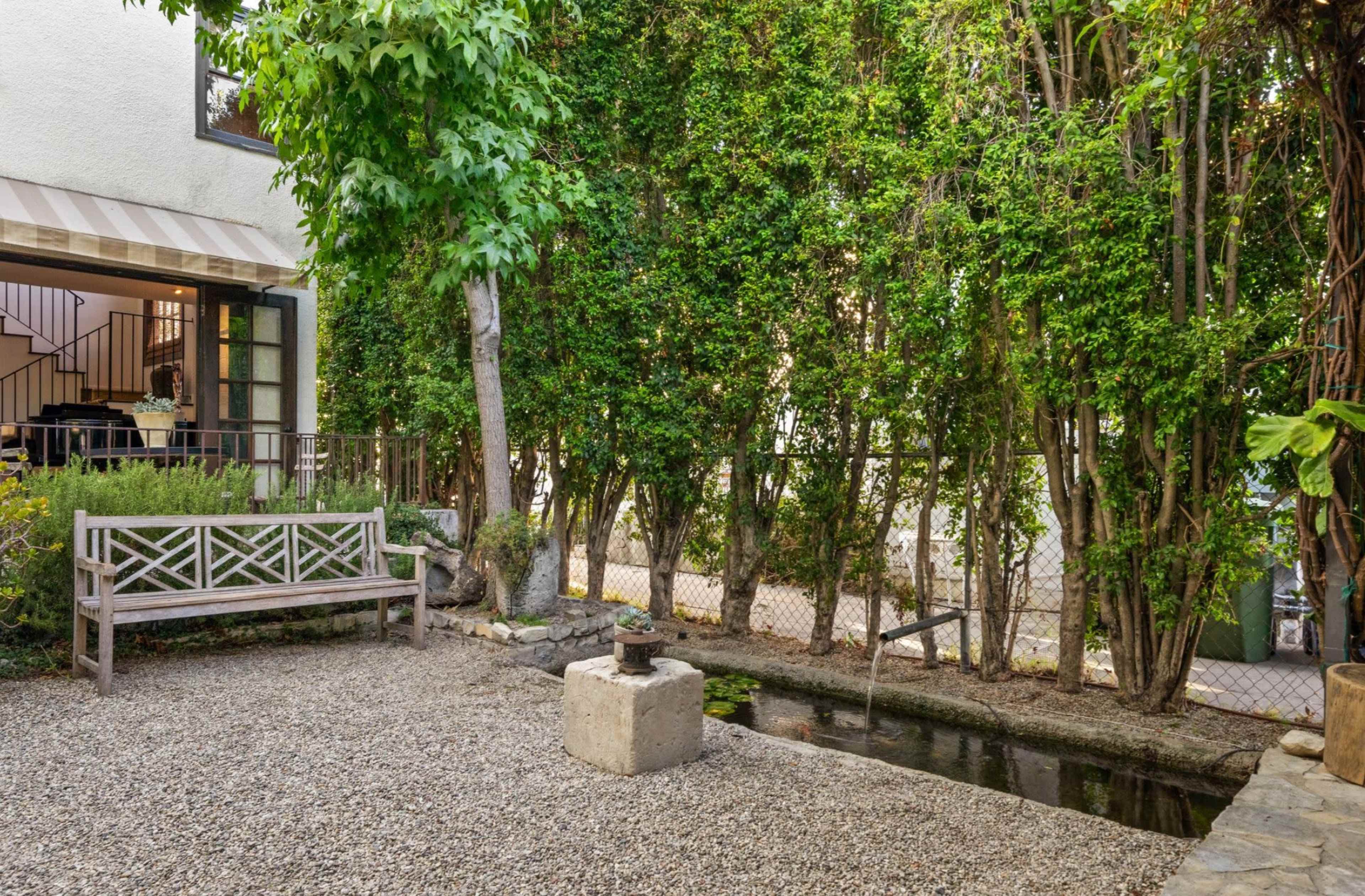 The image depicts a peaceful outdoor space featuring a gravel path, a wooden bench, and a small water feature surrounded by tall greenery.