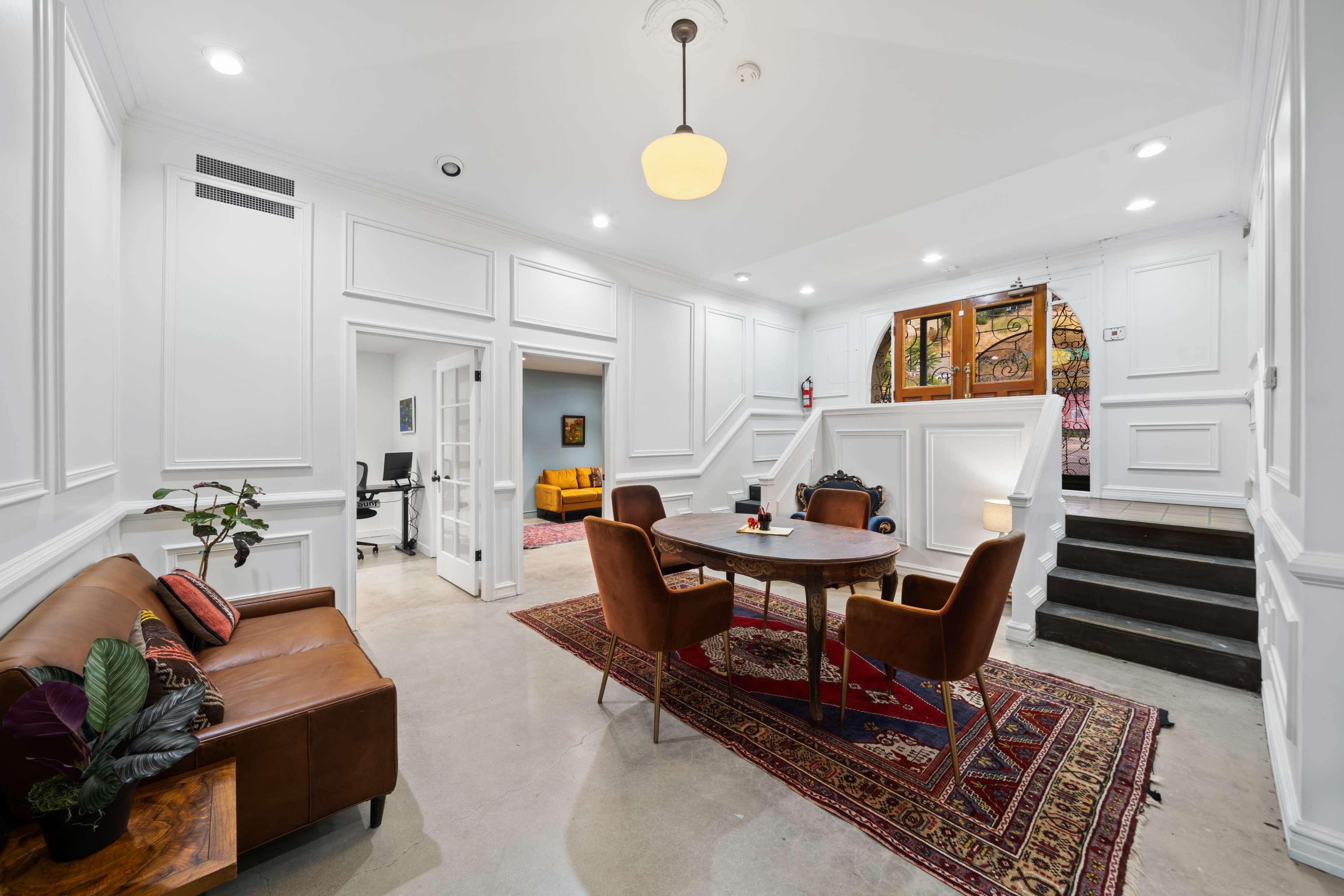 The image shows a modern interior space featuring a seating area with a brown leather sofa and a round table surrounded by brown chairs, leading to a staircase and rooms with artwork on the walls.