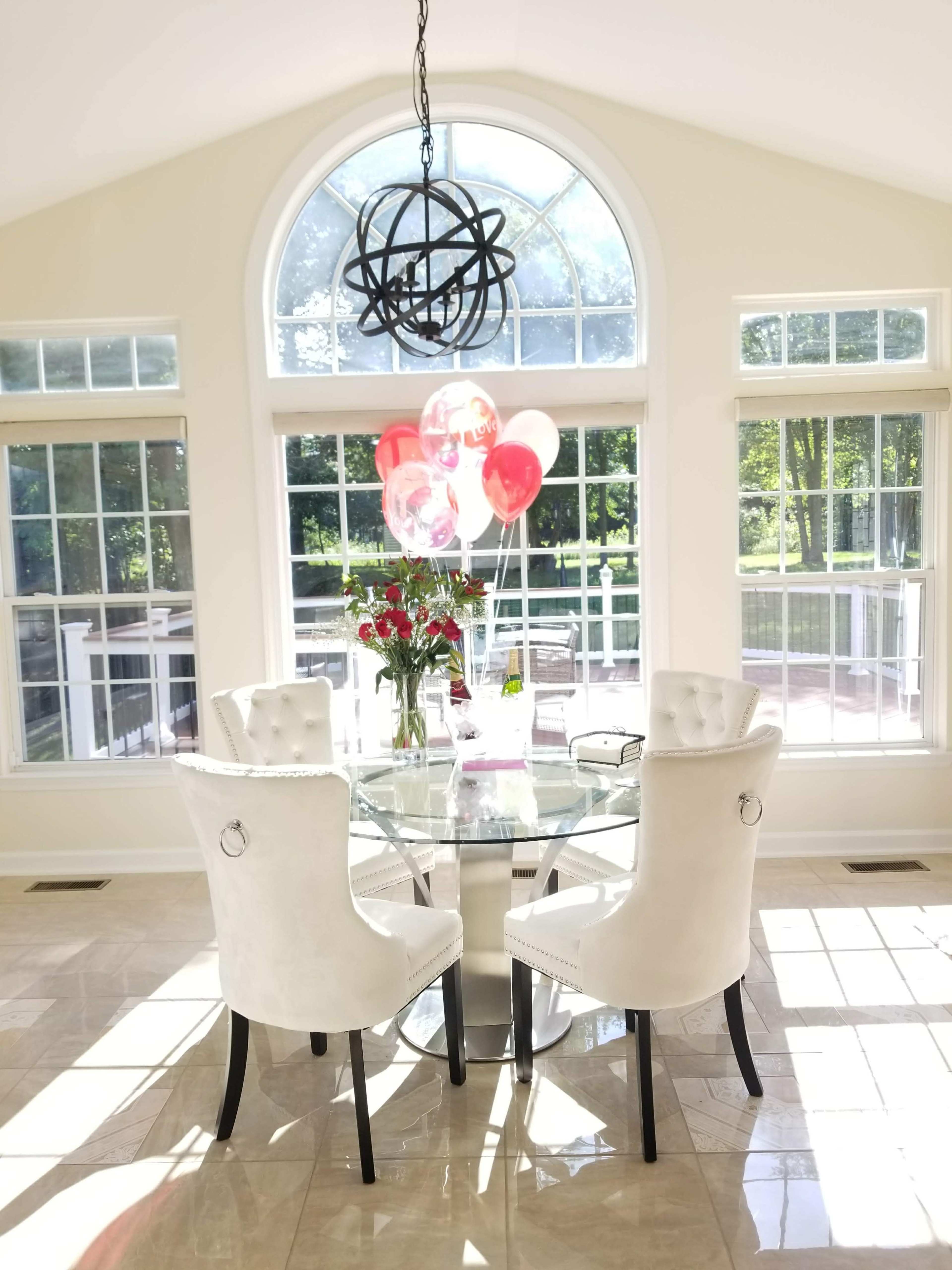 A round glass dining table with two white upholstered chairs is set against a backdrop of large windows, featuring a bouquet of roses and several balloons.