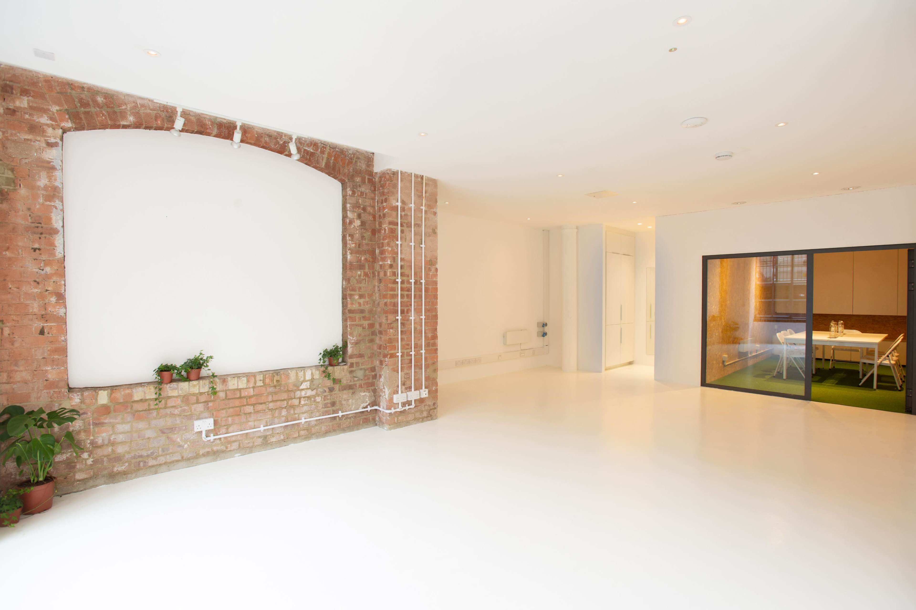 A spacious room with exposed brick walls, a white floor, and a large glass door leading to a kitchen area.