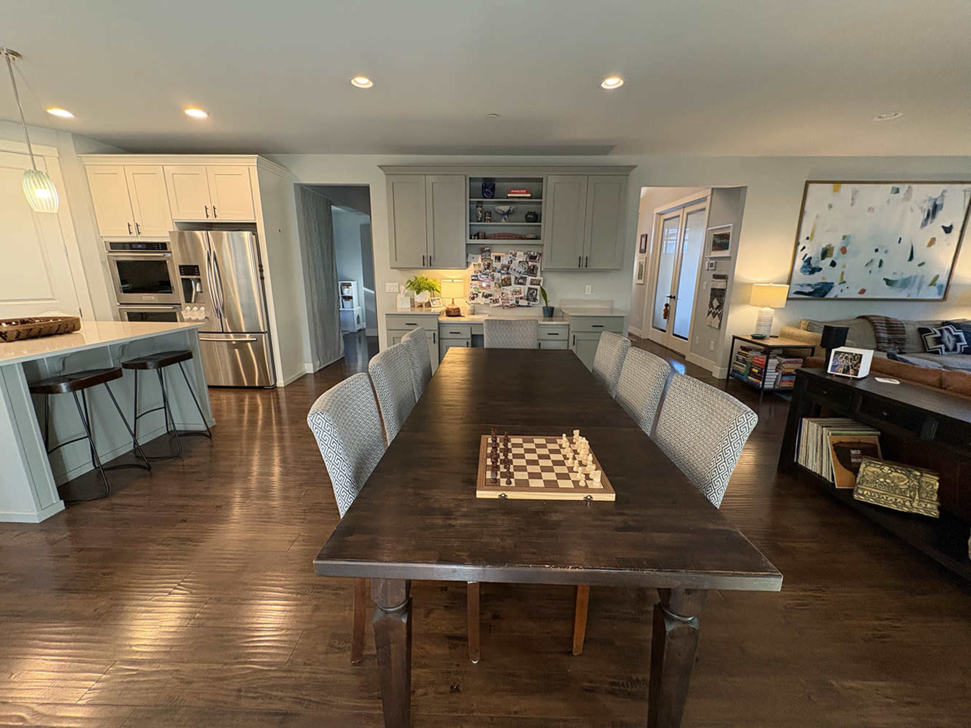 A large dining table with a chessboard is set in a spacious, modern kitchen and dining area, featuring light cabinetry and hardwood floors.
