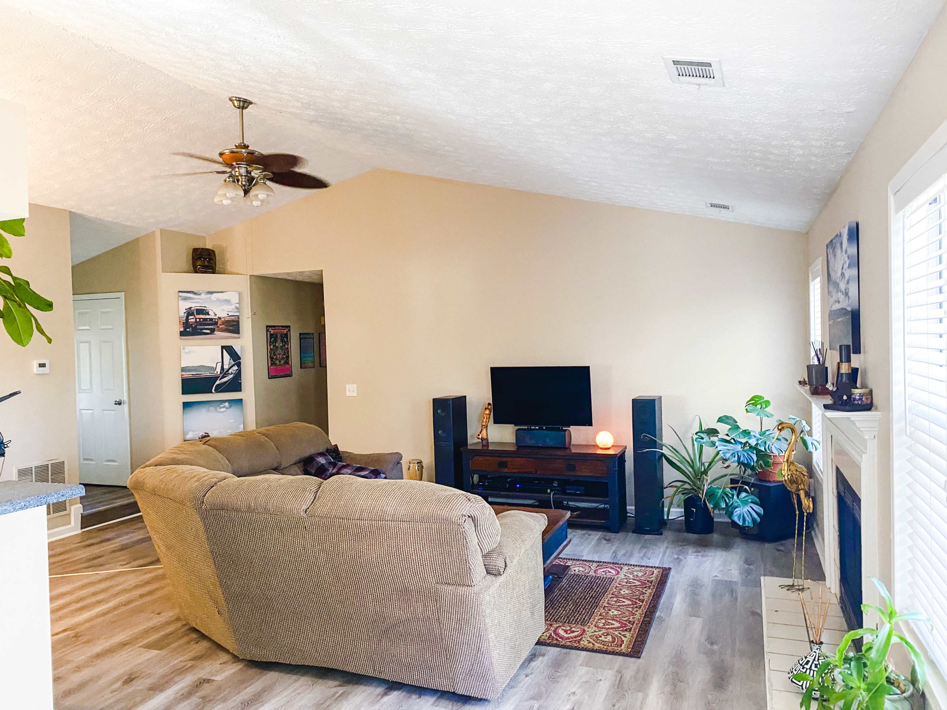 The living room features a large sectional sofa, a television stand with a TV, and houseplants, all set against light-colored walls and a ceiling fan.