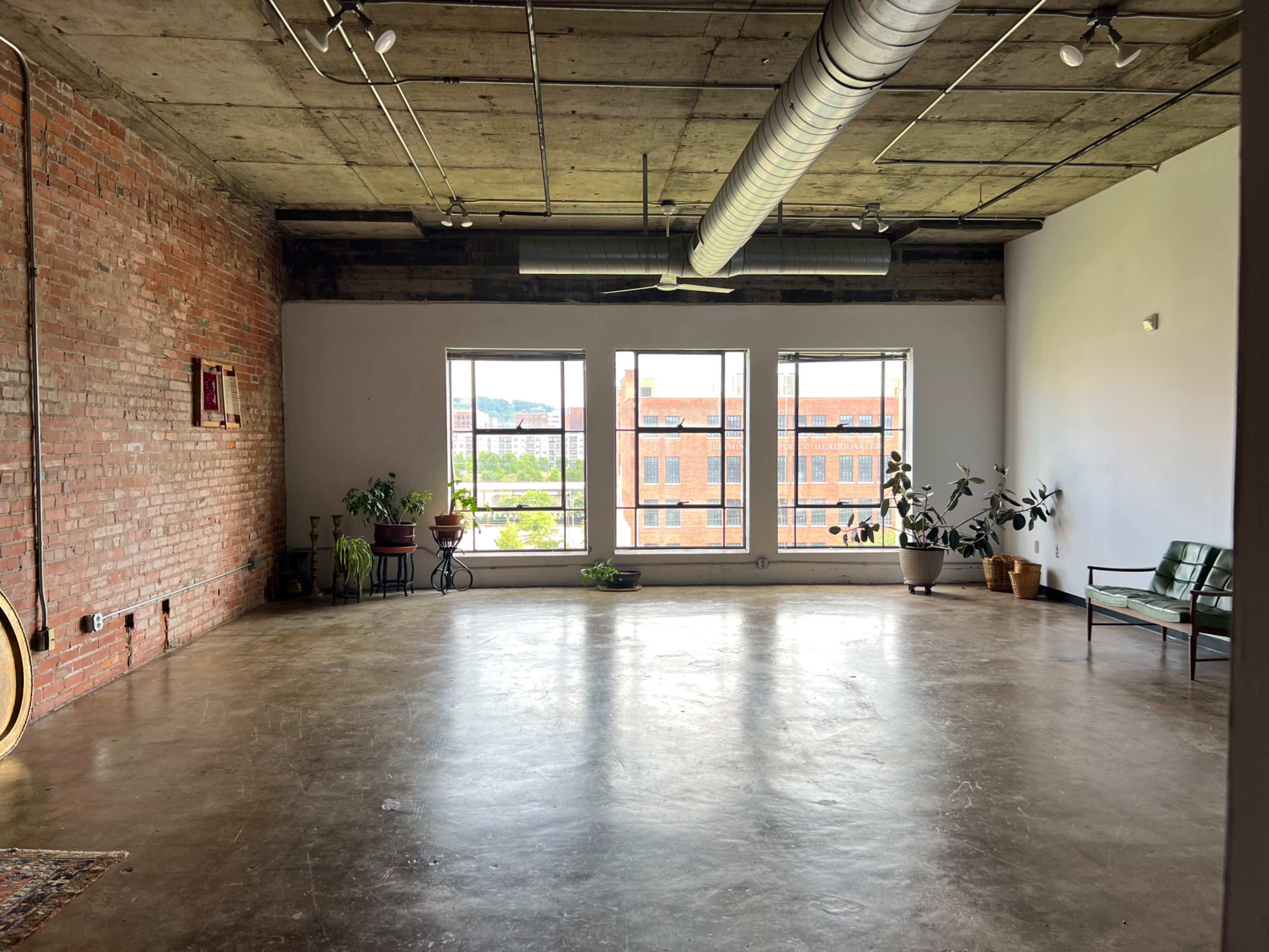 A spacious room with brick walls, large windows, and a concrete floor, furnished with a green sofa and several potted plants.