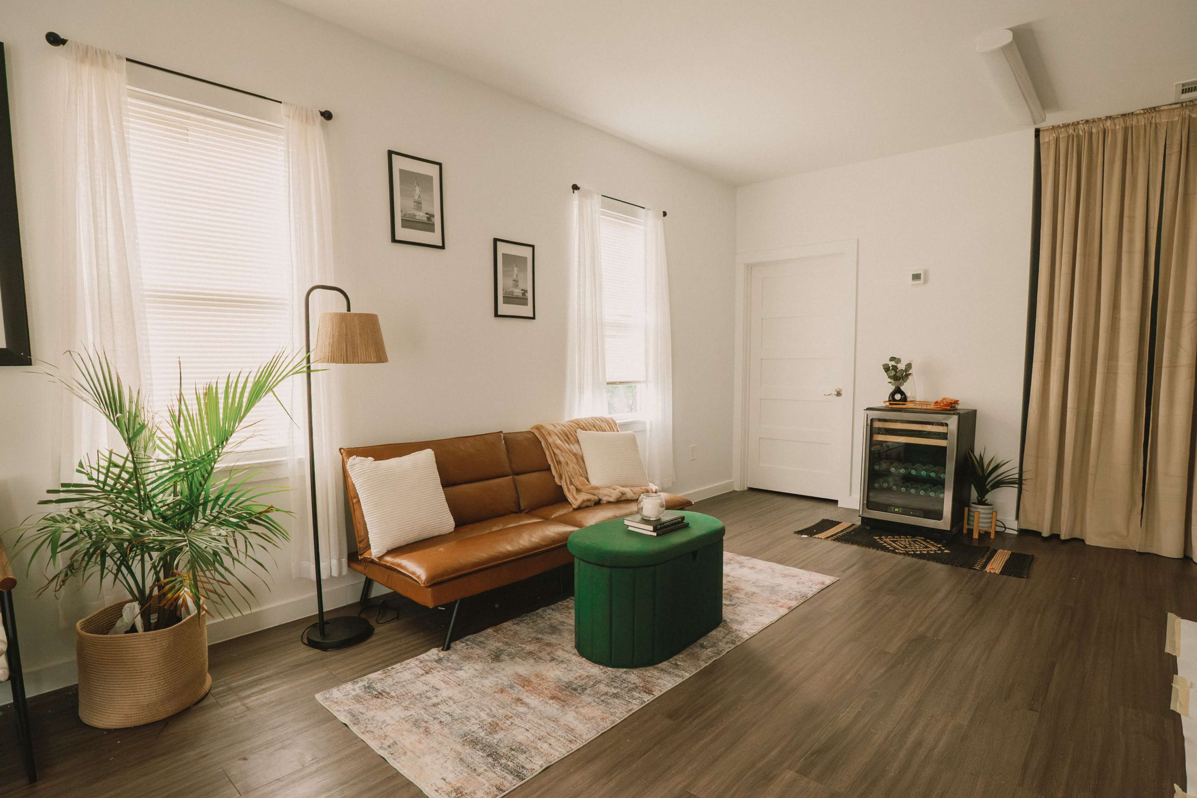 The room features a brown leather sofa, a green ottoman, a potted plant, and a small beverage refrigerator, with light streaming through the windows.