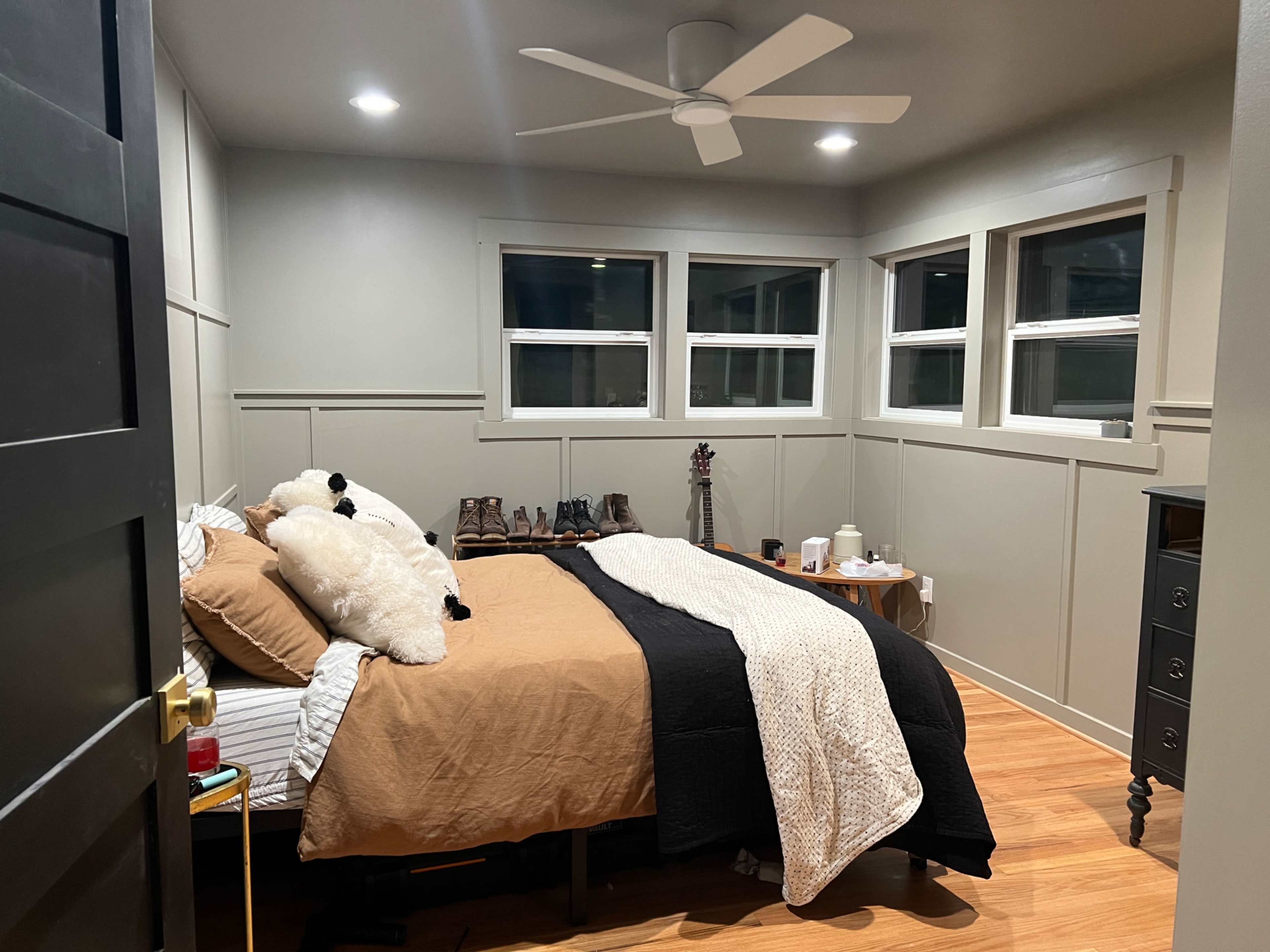The image shows a bedroom featuring a bed with brown and black bedding, wooden flooring, and several windows allowing light to enter.