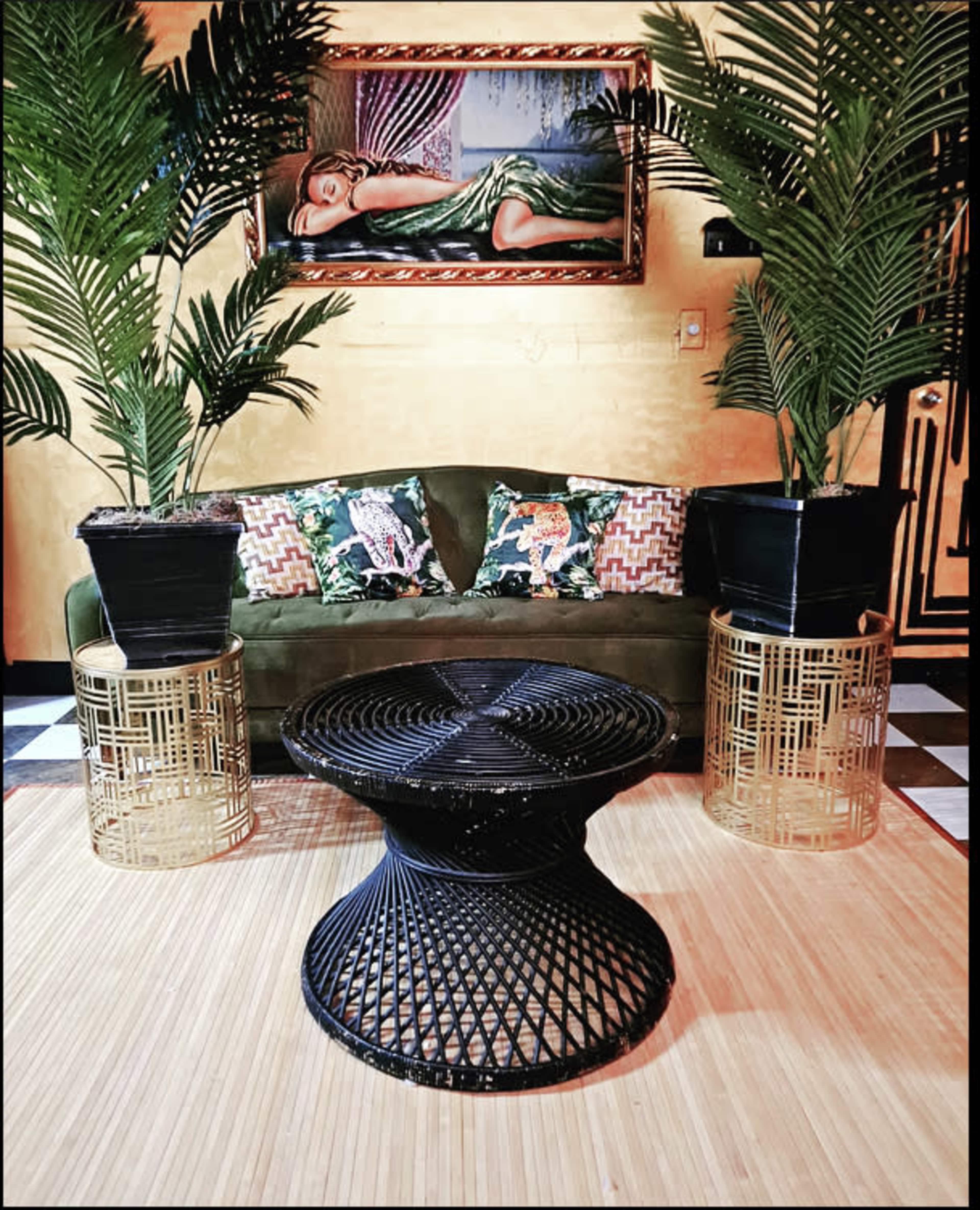 A green sofa with patterned cushions is positioned in front of a round black coffee table, flanked by tall potted palm plants against a backdrop featuring a painting.