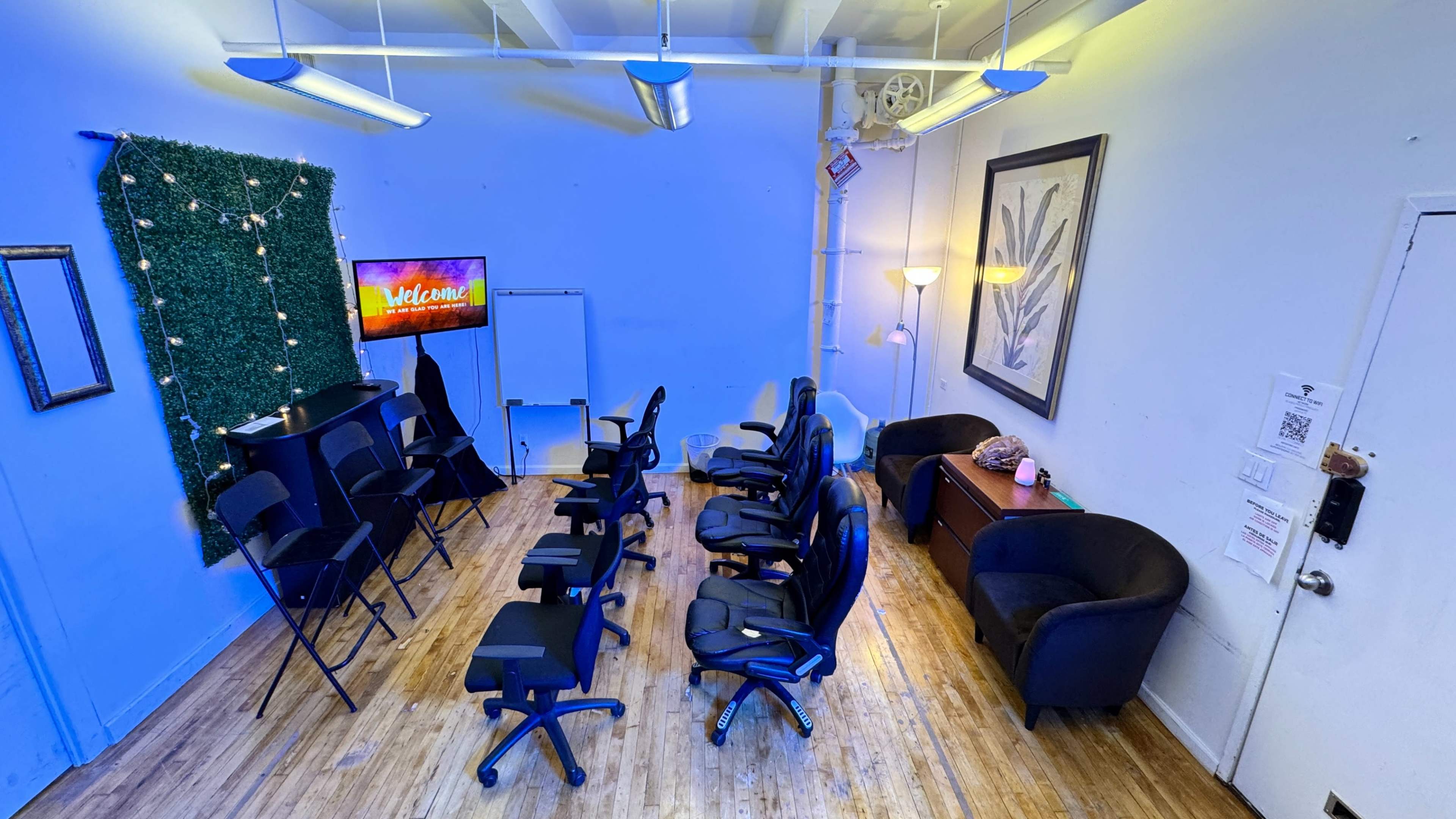 A room arranged with several black chairs, a television displaying a welcome message, and a green wall adorned with string lights.