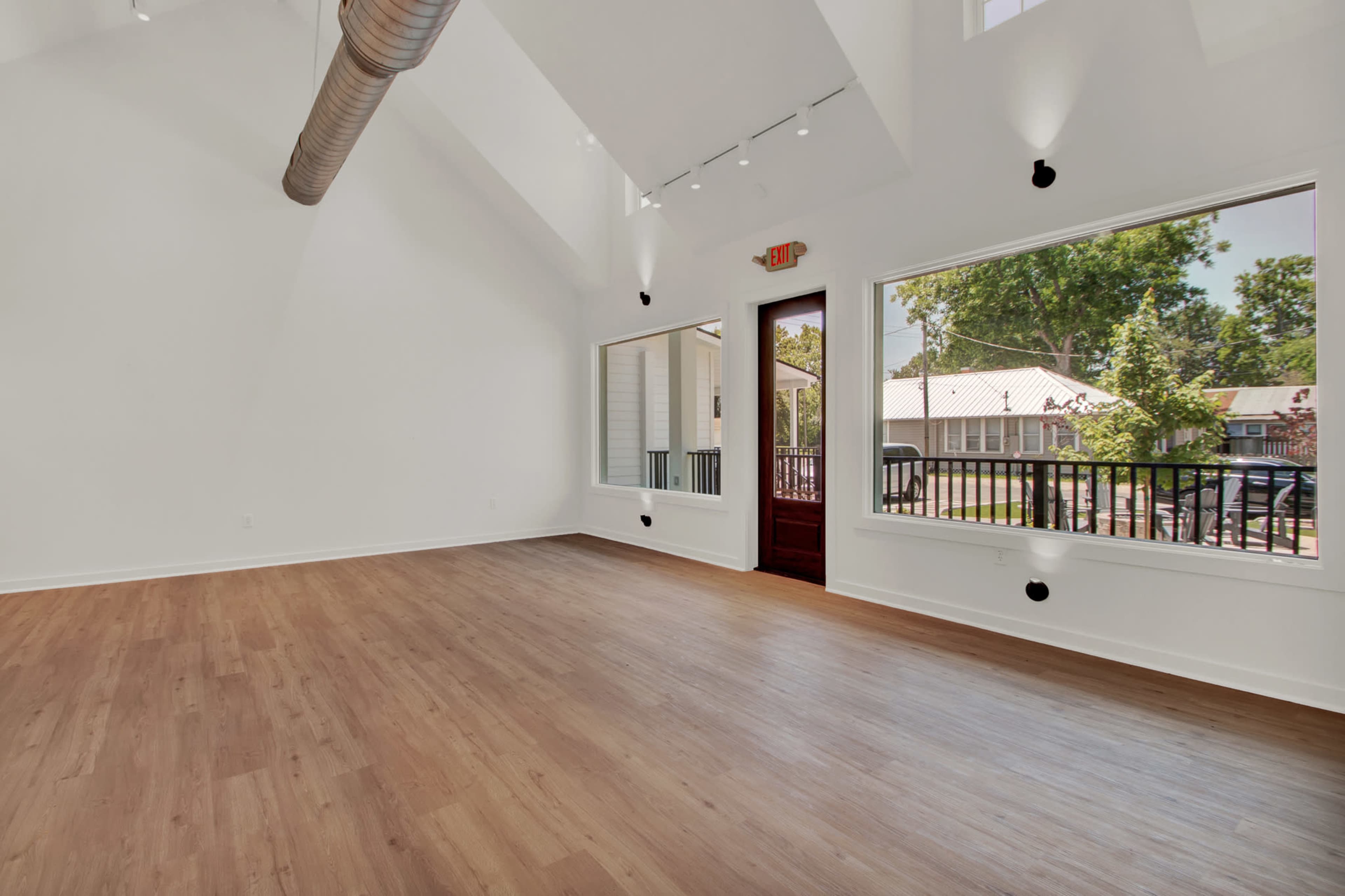The image shows a spacious, empty room with hardwood flooring, large windows letting in natural light, and a door leading to an outdoor area.