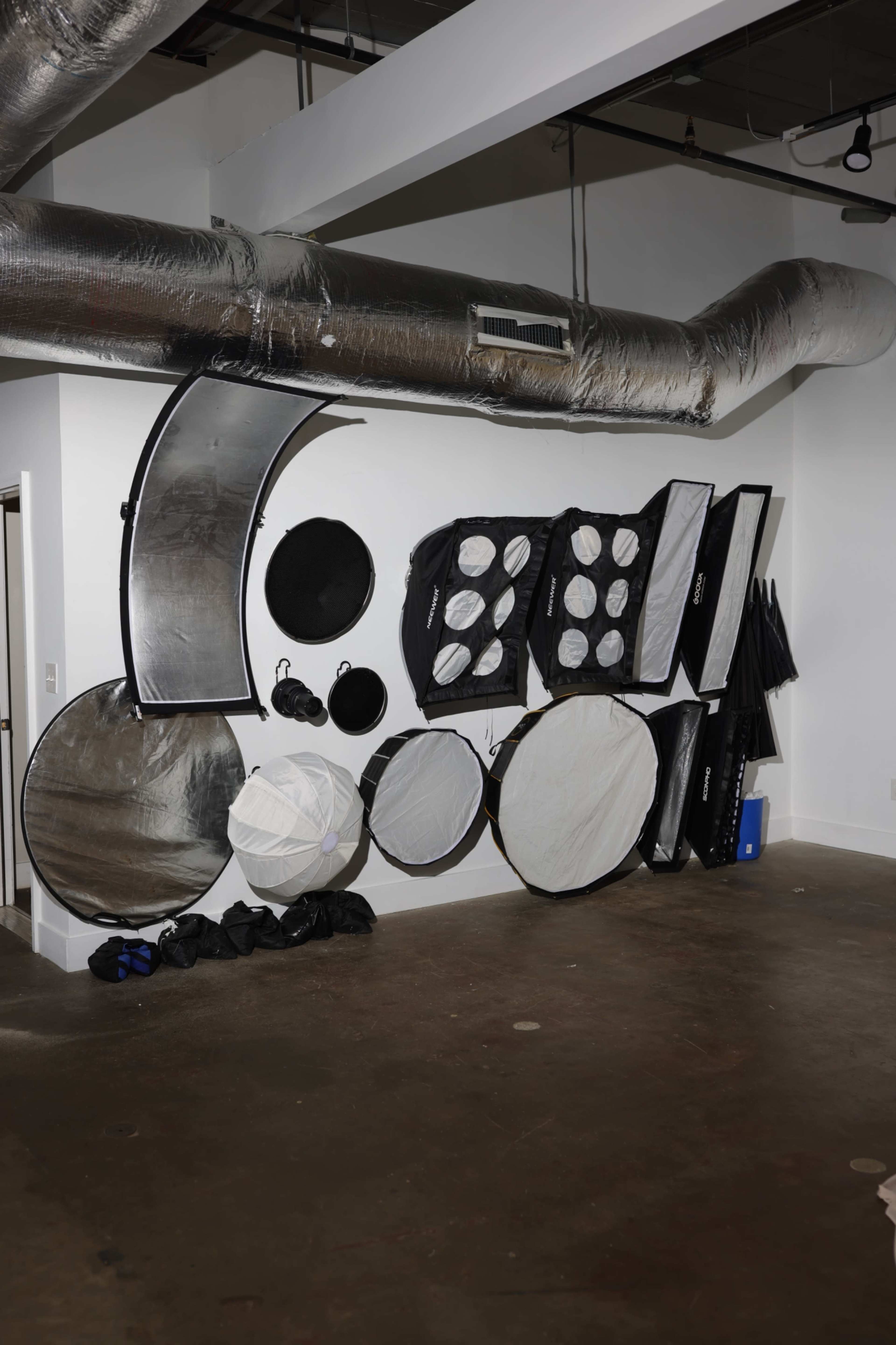 The image shows a wall with various photography light modifiers and reflectors arranged neatly, alongside some bags on the floor.