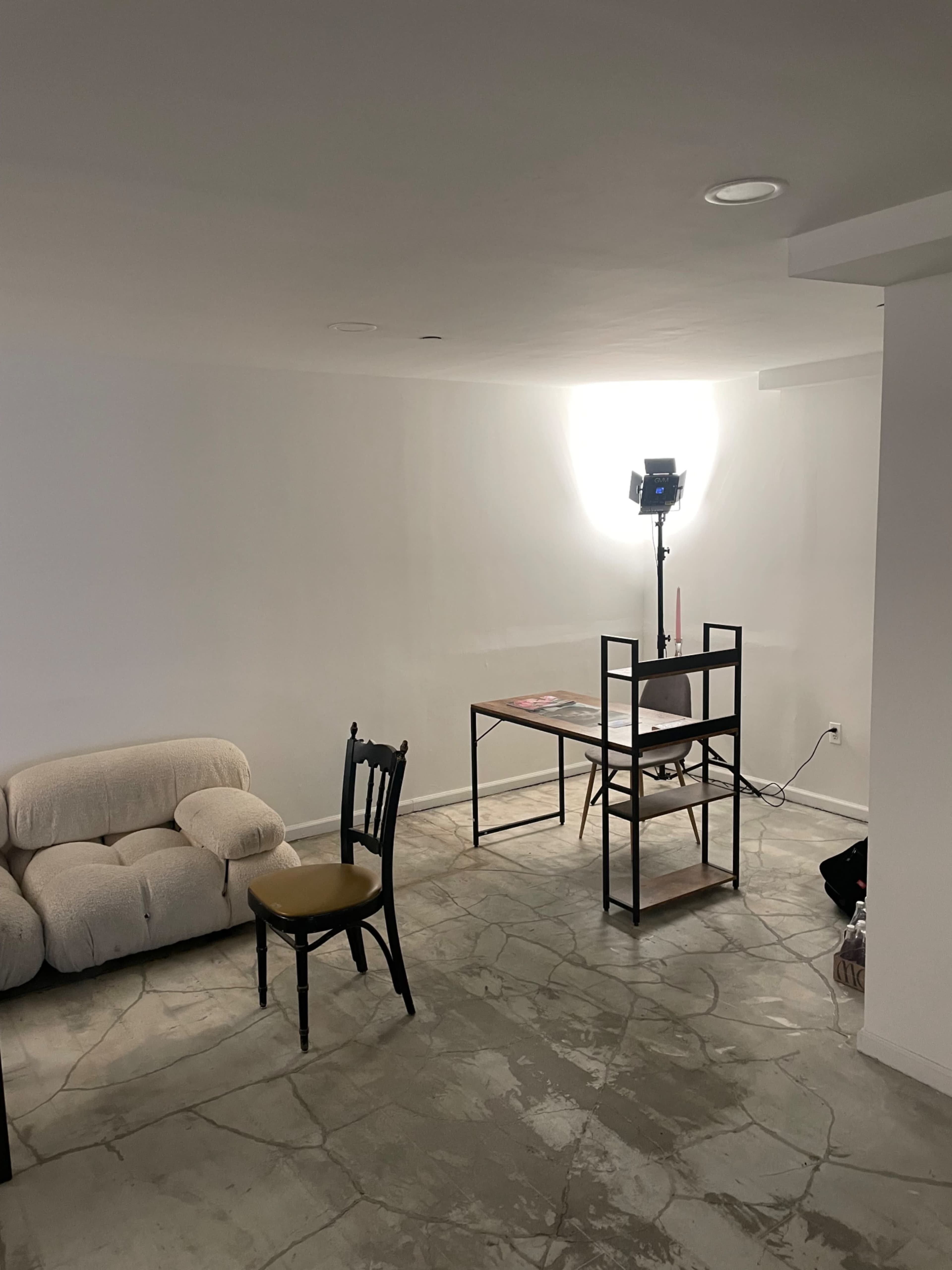 A minimalistic room features a beige couch, a black chair with a yellow cushion, a table with magazines, and a lighting setup in the background.