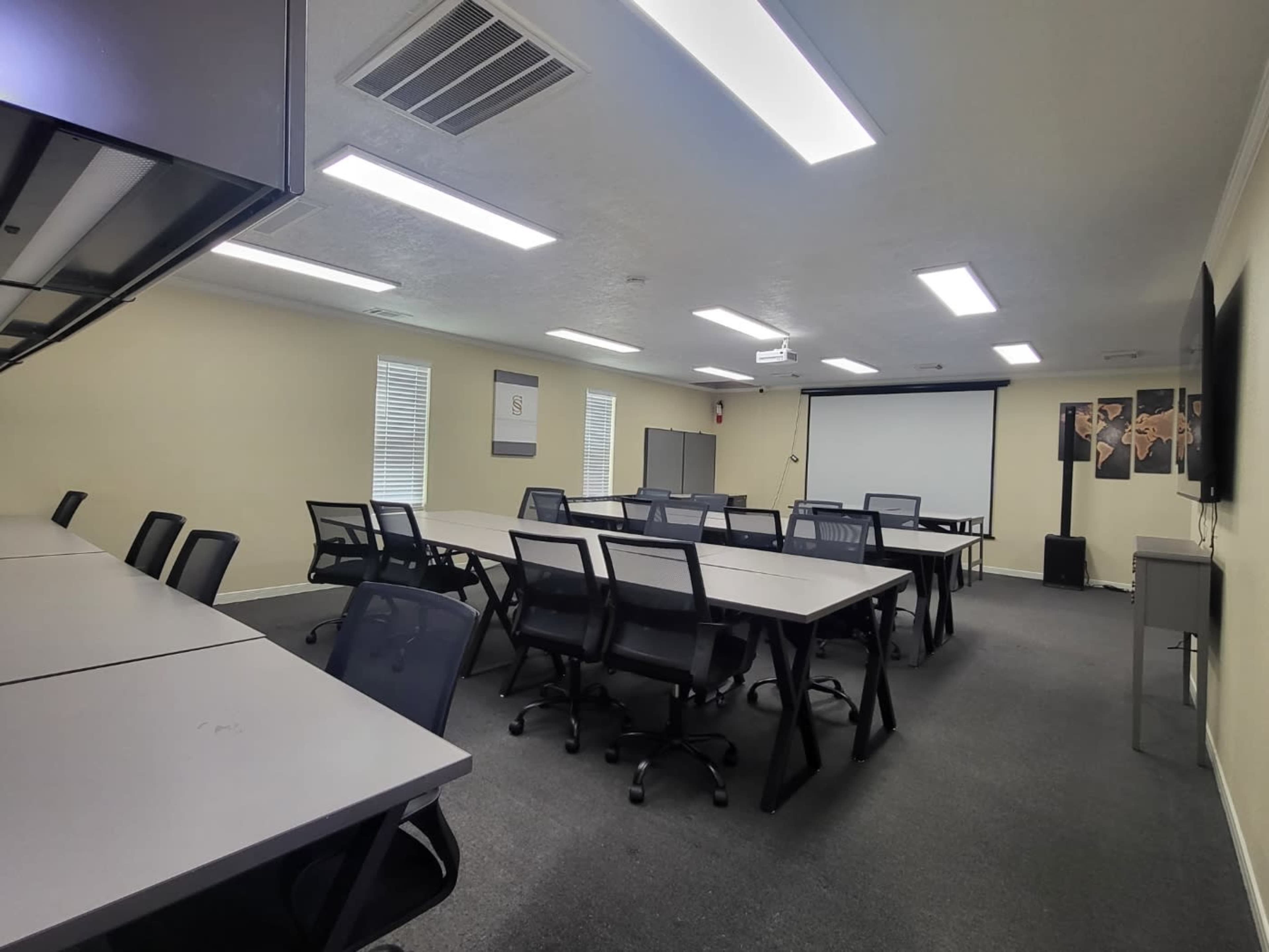 A spacious classroom features several rows of tables and chairs, a projector screen, and various educational materials on the walls.