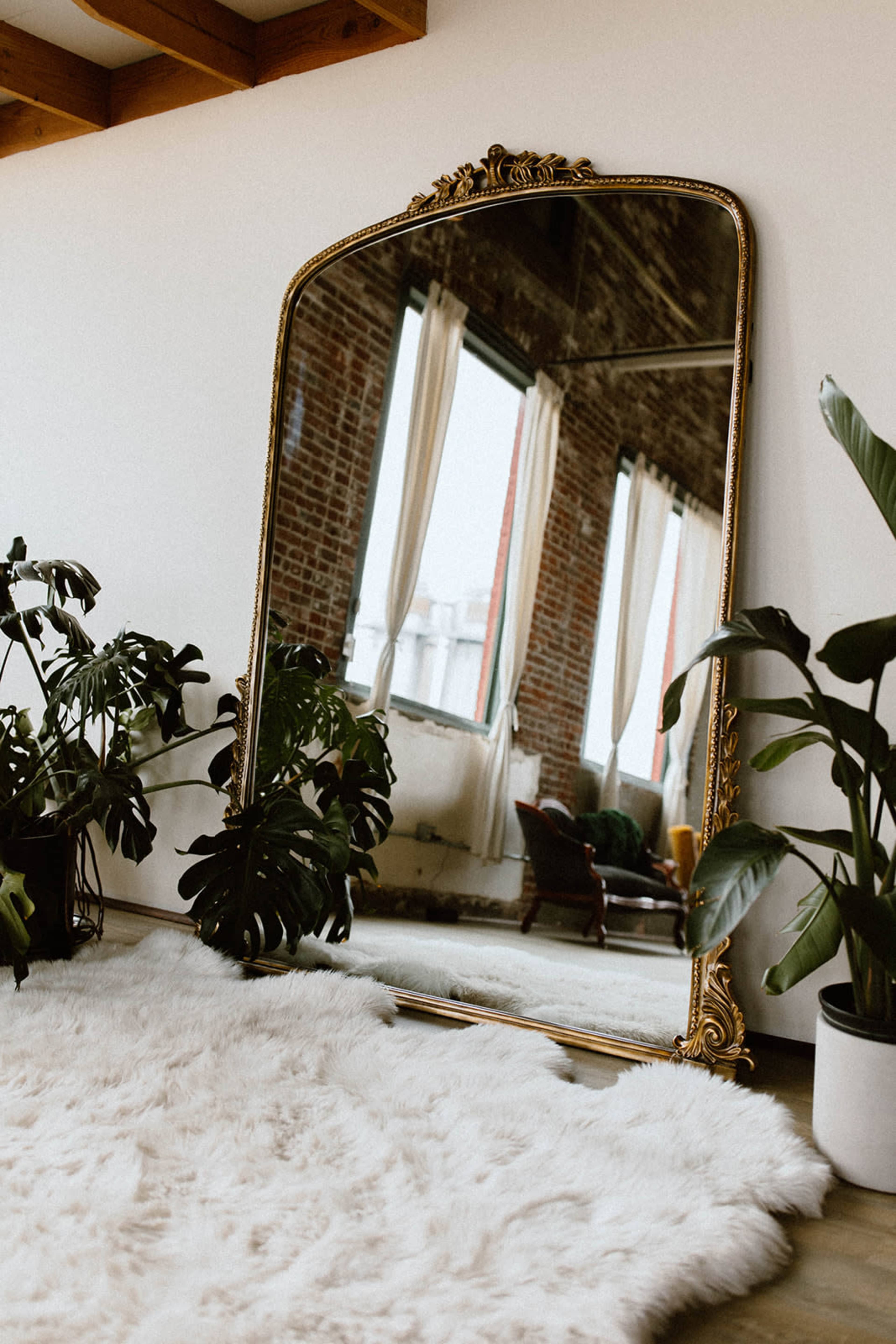 A large, ornate mirror stands against a wall, reflecting the interior of a room with plants and a cozy seating area.