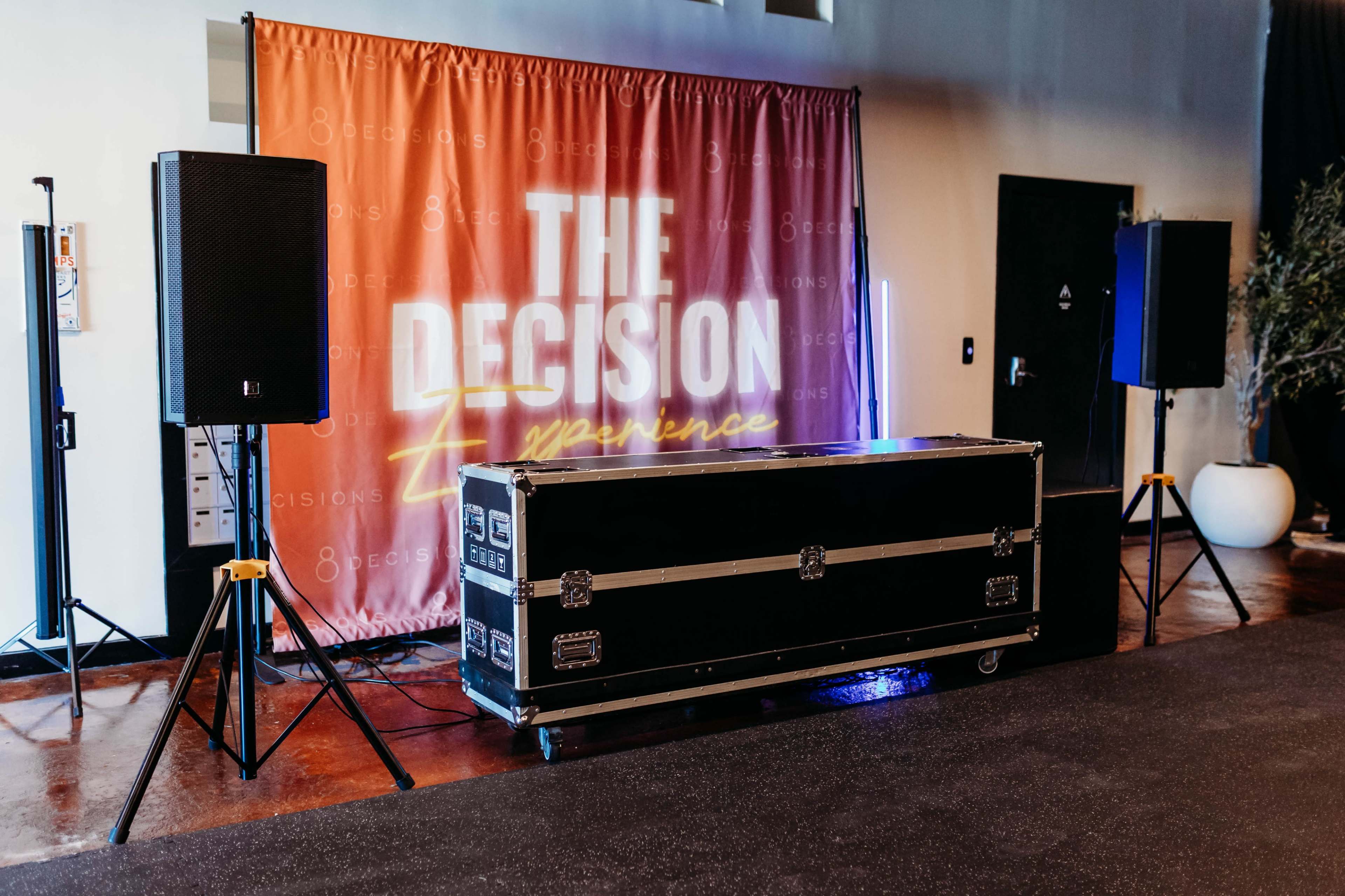 The image shows a stage area with a backdrop that reads "THE DECISION Experience," flanked by speakers and a large black trunk on wheels.