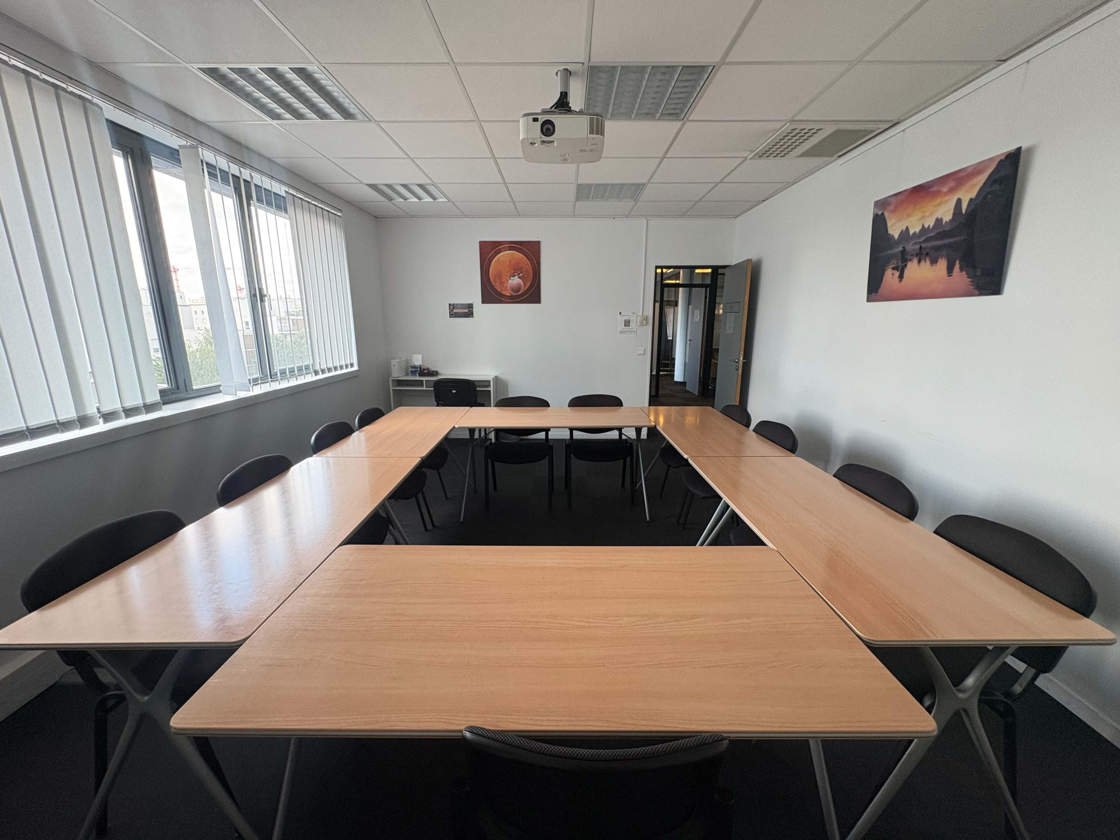 The image shows a spacious conference room with a large U-shaped arrangement of tables and black chairs, large windows, and a projector mounted on the ceiling.