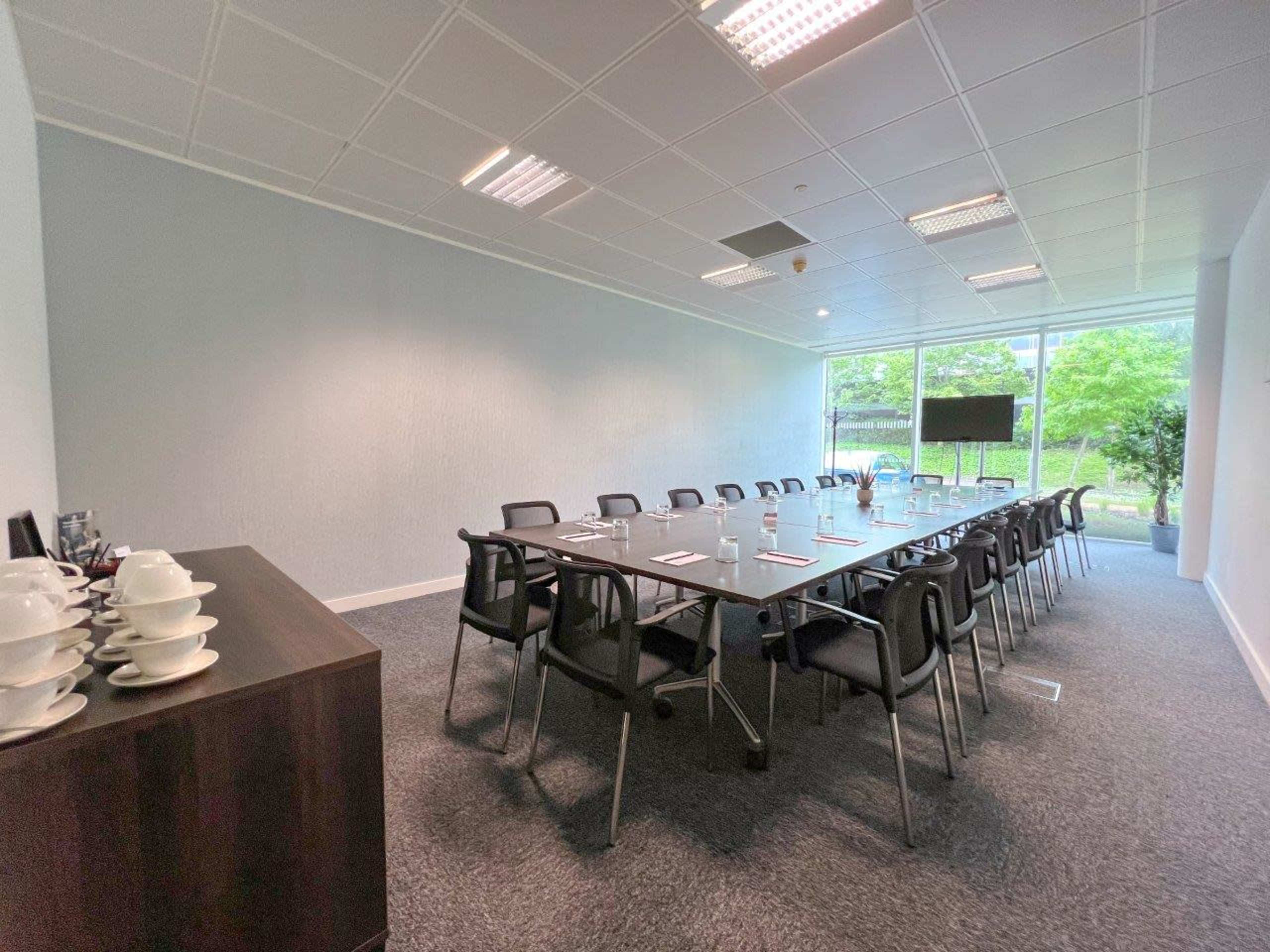 A conference room is set up with a long table surrounded by chairs, with a large screen and windows overlooking greenery.