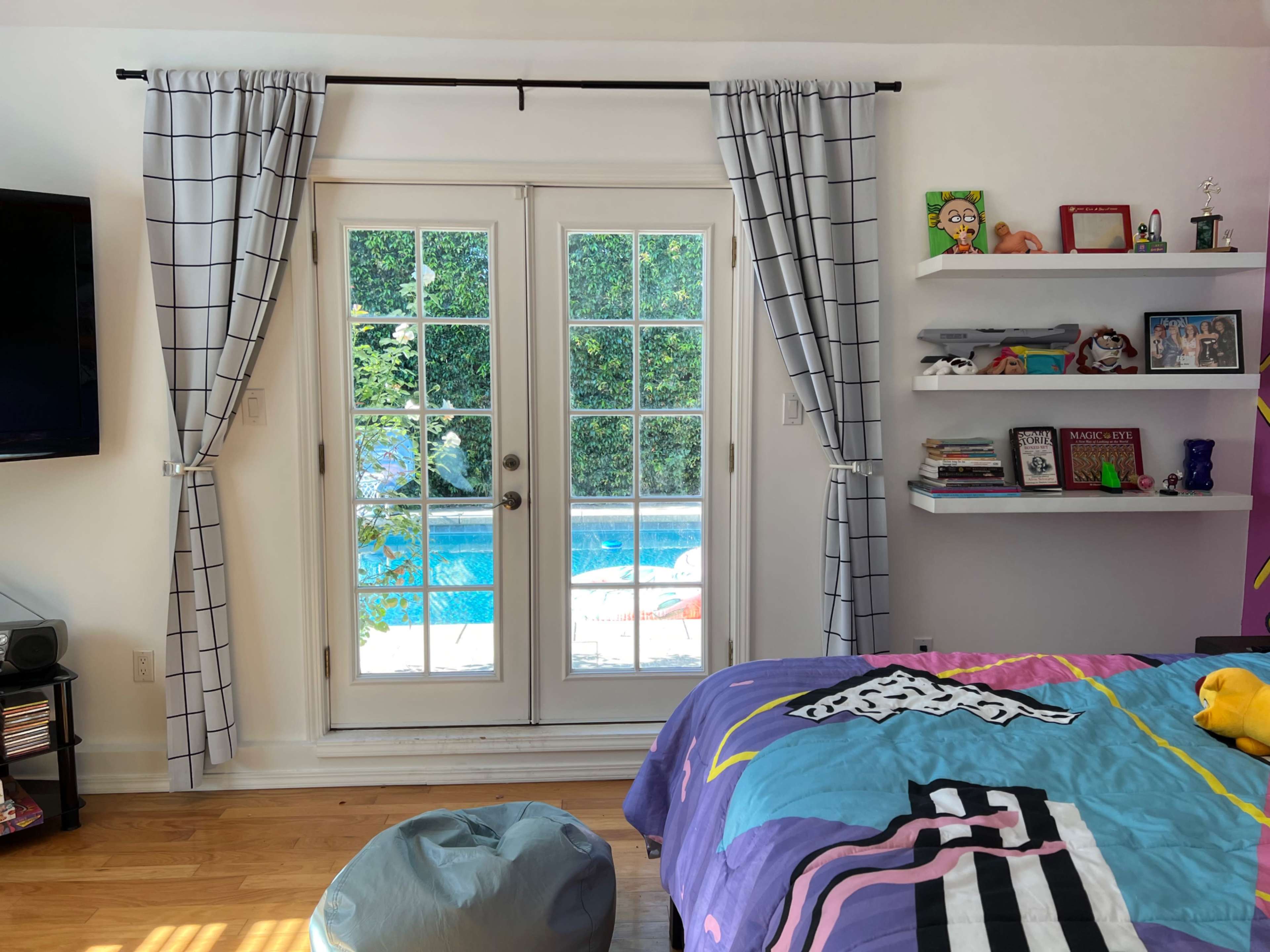 A brightly colored bedroom with a bed covered in a patterned blanket, a window with sheer curtains, and a view of a pool outside through glass doors.