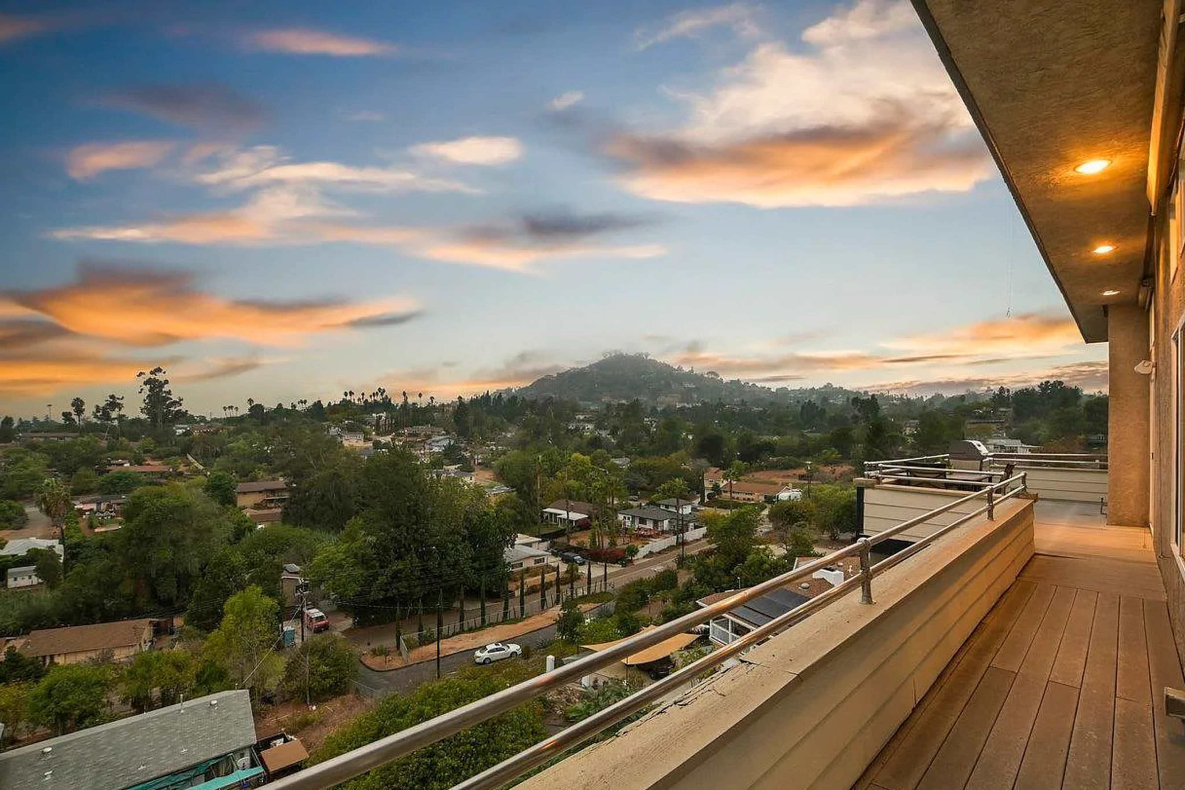 A balcony view overlooking a residential area with houses and trees, set against a backdrop of hills and a colorful sky at sunset.