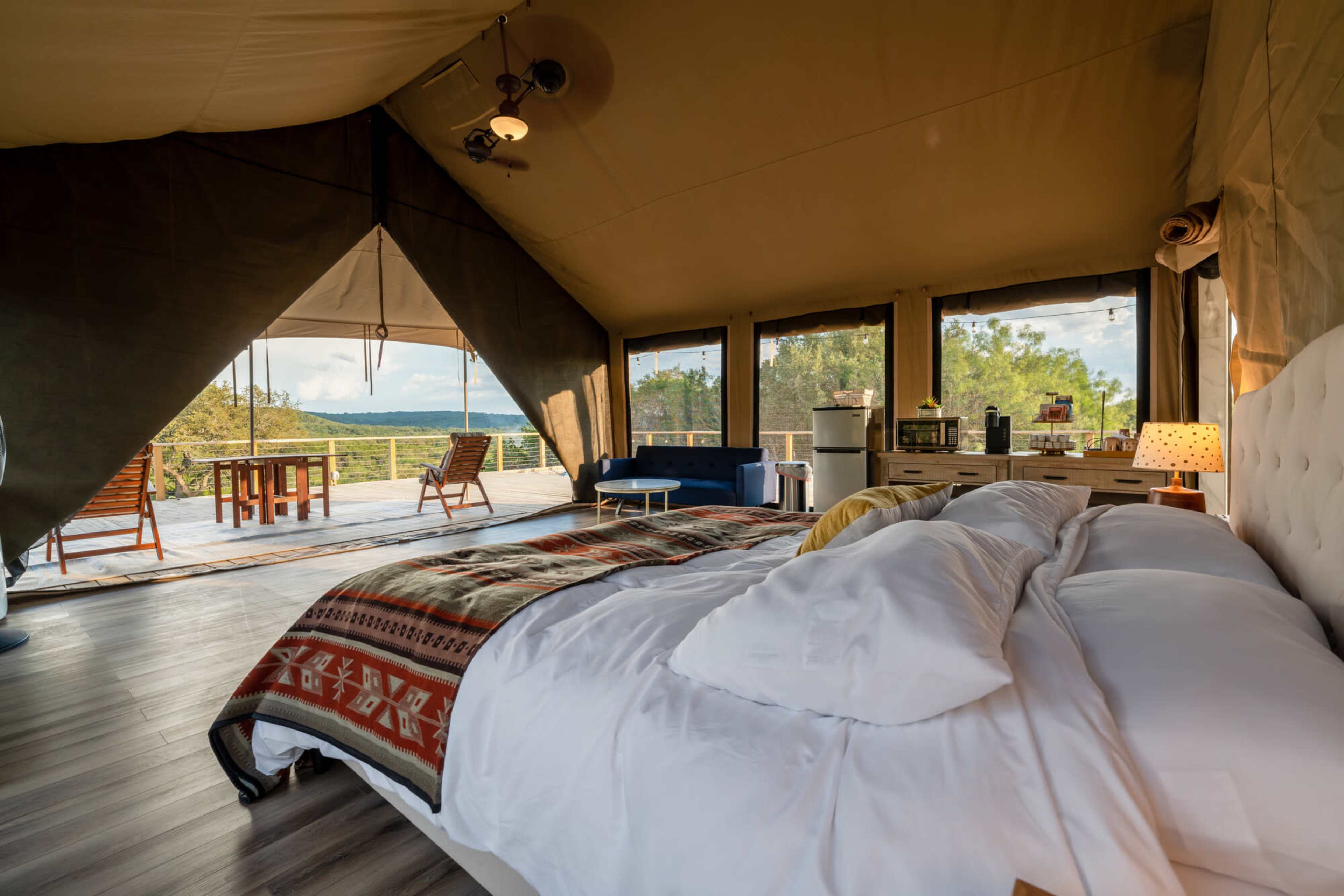 The image shows a spacious glamping tent interior with a comfortable bed, a seating area, and large windows overlooking a scenic outdoor deck.