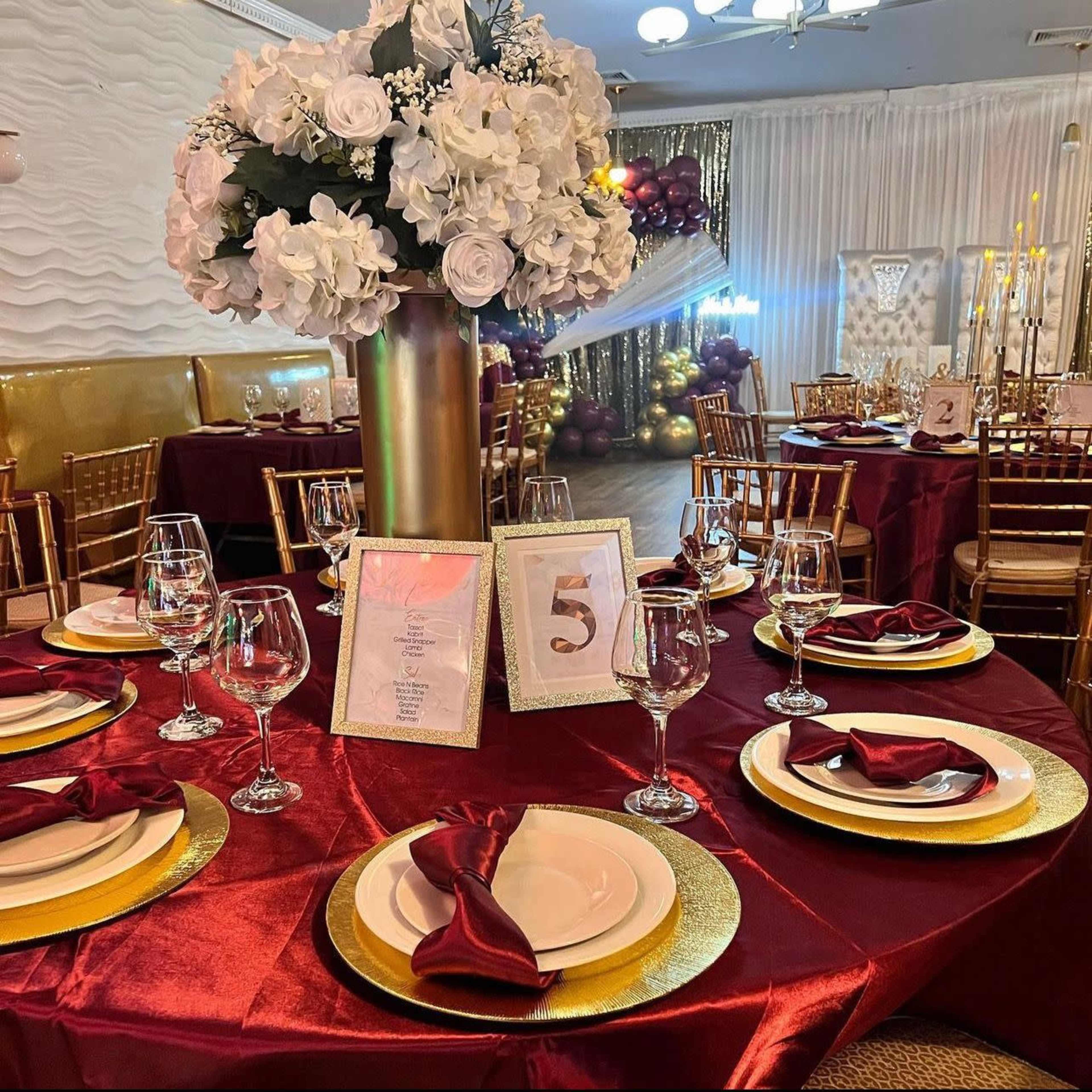 A banquet table is set with elegant red and gold decorations, including glassware, white plates, and a large floral centerpiece.