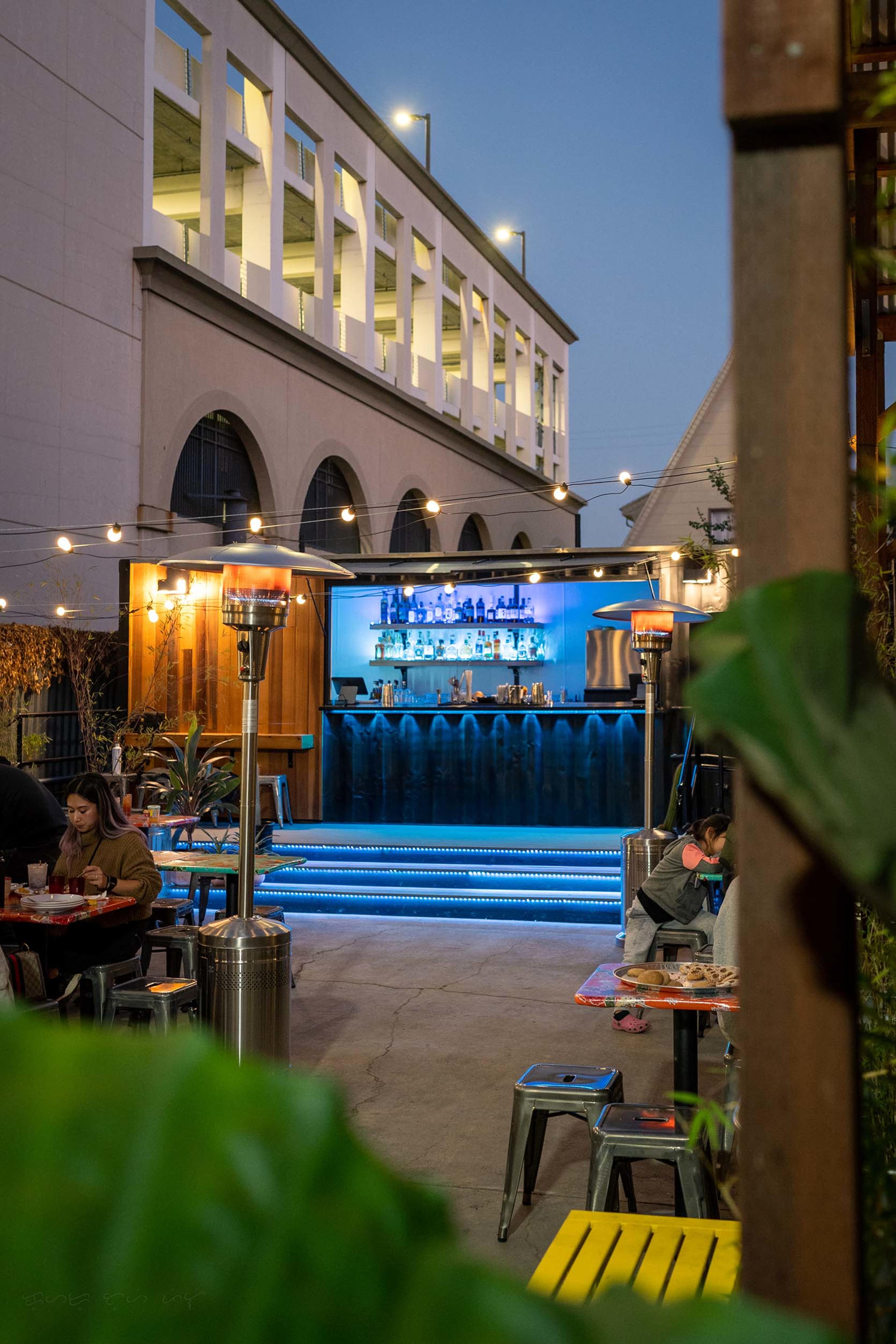 A courtyard bar features illuminated shelves of bottles behind a water wall, with seated patrons and string lights overhead.