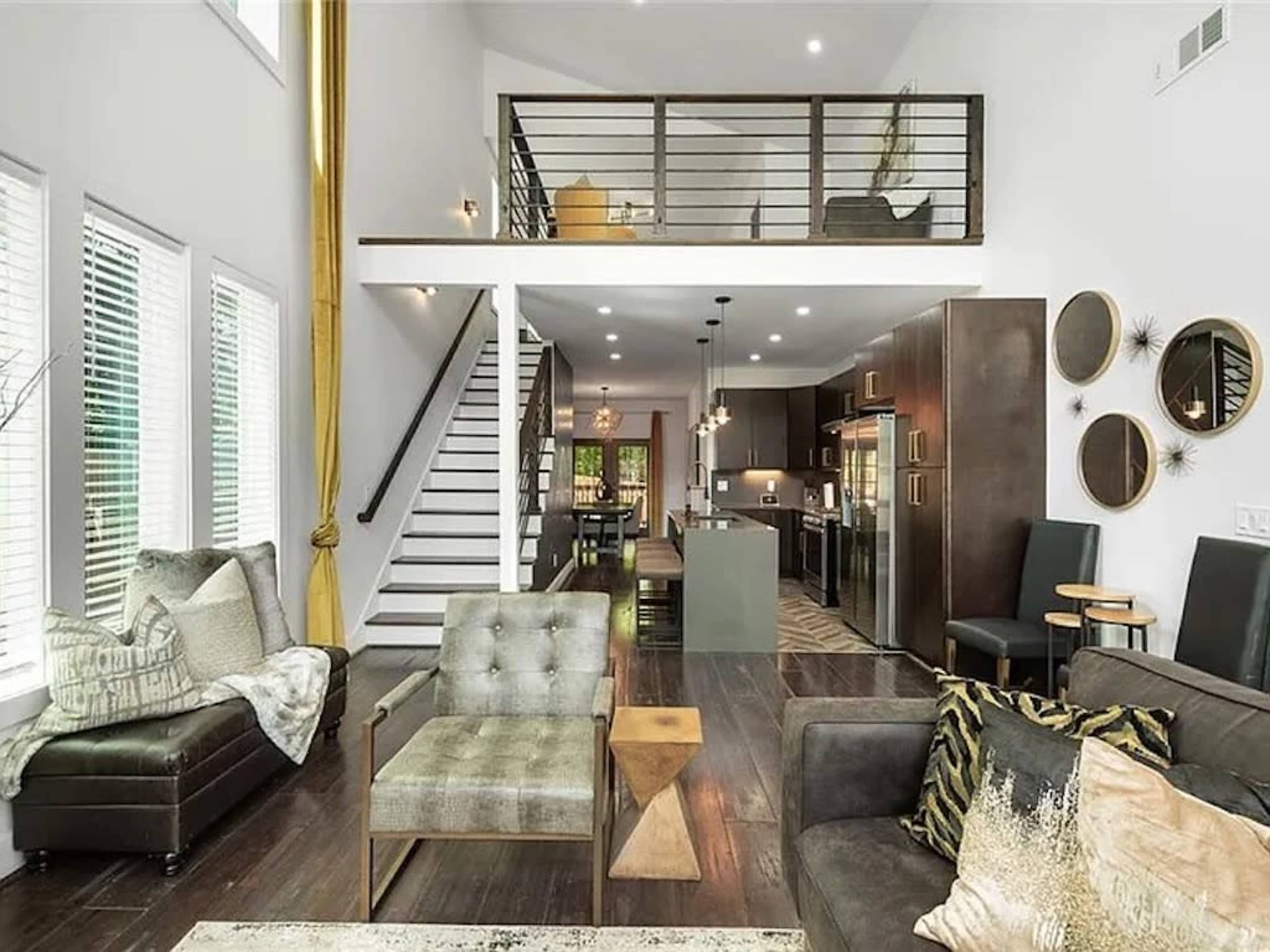 A modern living space featuring a mix of seating areas, a staircase, and a kitchen, all illuminated by natural light through large windows.