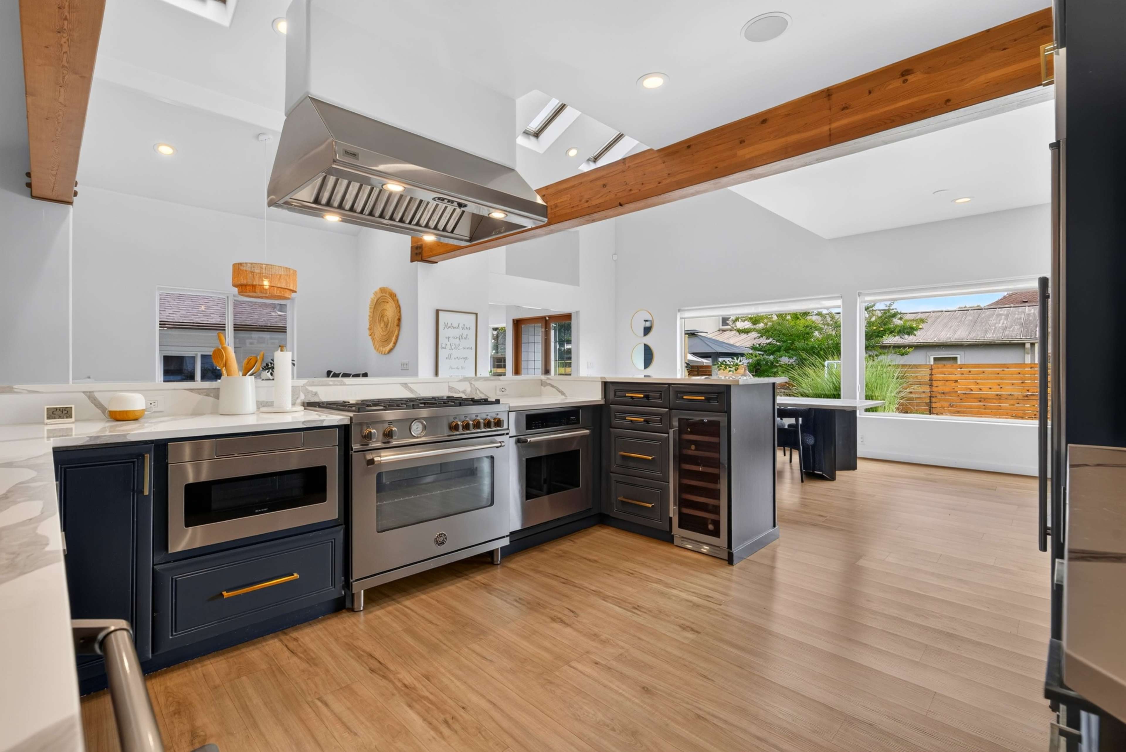 The image shows a modern kitchen featuring stainless steel appliances, dark blue cabinets, and wooden beams, with large windows providing natural light.
