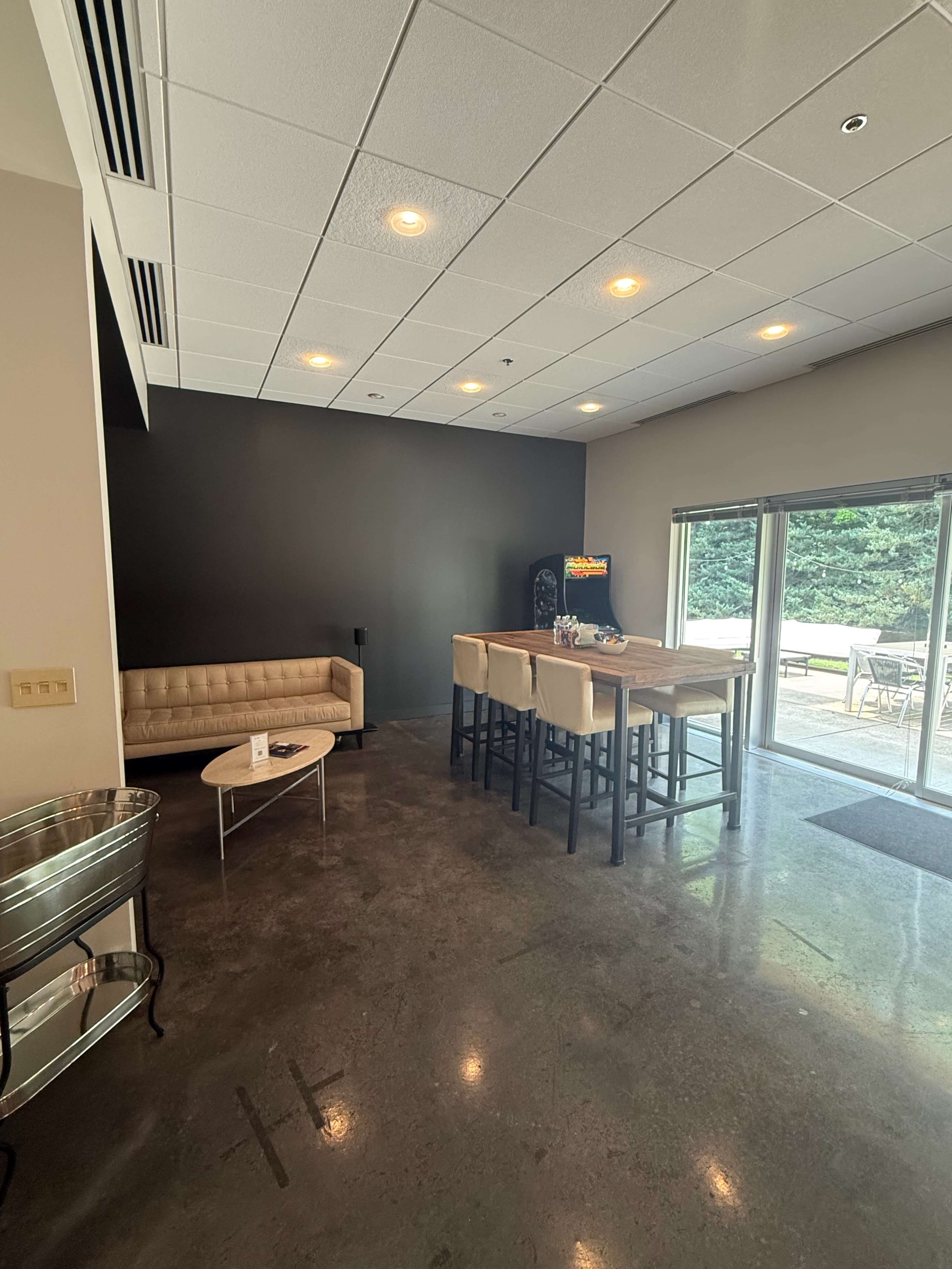 The image shows a modern lounge area with a beige couch, a small coffee table, and a bar-height table with chairs, alongside large sliding glass doors that lead to an outdoor patio.