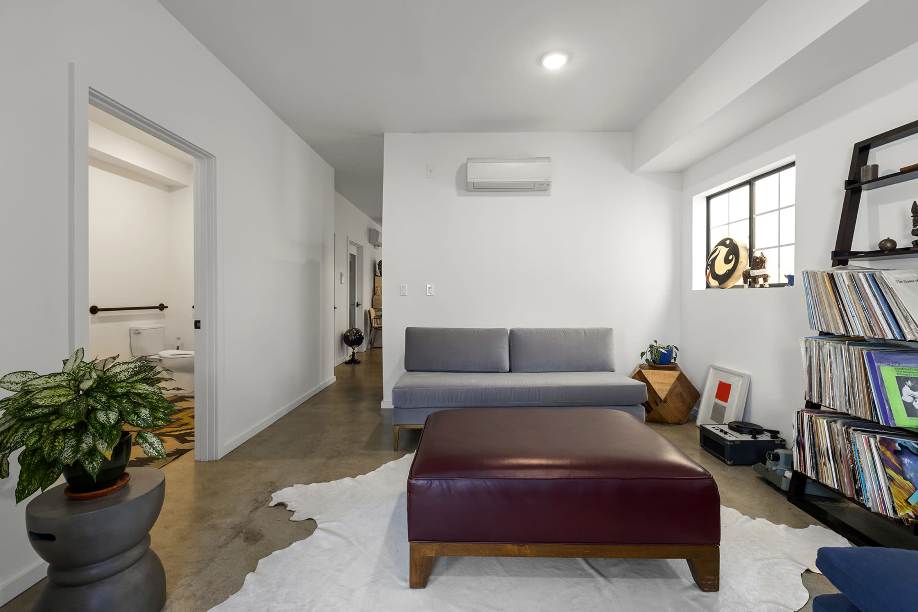 A modern living room features a gray sofa, a maroon ottoman, and a collection of vinyl records displayed on a shelf against a white wall.